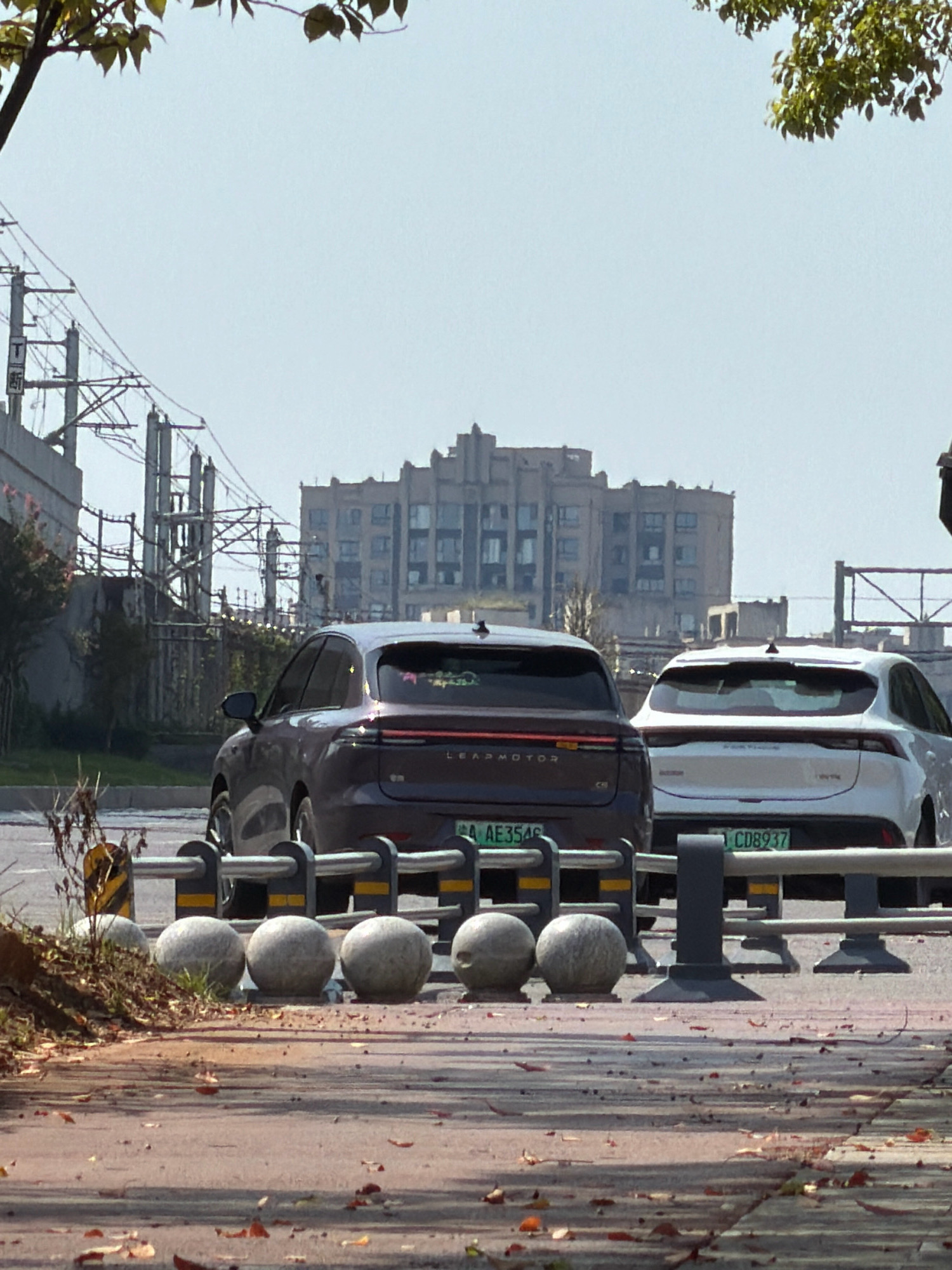 The image captures a street scene in the city of Jiangjin, China, on a clear, sunny day. In the foreground, a paved walkway with scattered autumn leaves is visible. Separating the walkway from the street is a row of spherical concrete bollards. The street itself features two cars parked side-by-side. The car on the left is a dark-colored sedan with a license plate that reads "AE3546". The car on the right is a white hatchback with a license plate that reads "CD8937". Both cars appear to be stationary.
In the background, industrial infrastructure, including electrical poles and wires, suggests an urban or semi-industrial area. Beyond this, a large, multi-story building with a distinct architectural style, possibly aged or historical, dominates the skyline. The sky is clear and light blue. The overall scene depicts a typical street view, with vehicles, infrastructure, and buildings characteristic of a developed city environment.