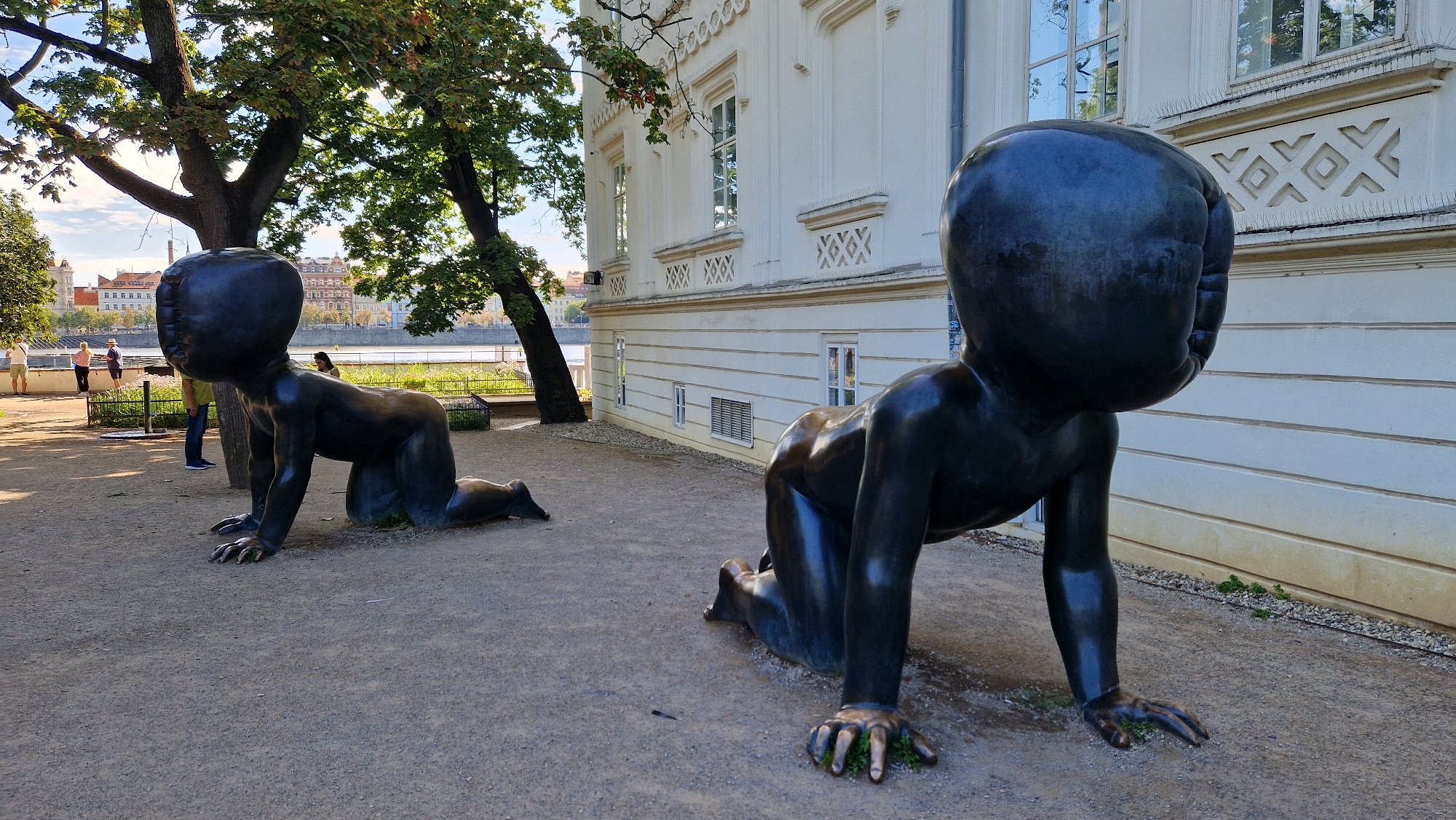 This image shows a scene in Prague, Czechia, featuring two large, dark, stylized sculptures of babies crawling. The sculptures are positioned on a gravelly ground surface, with one sculpture in the foreground and another further back and to the left. Both sculptures have large, featureless heads and appear to be made of a dark, metallic material.
Behind the sculptures, there is a large, white building with windows and decorative architectural elements. To the left of the building and sculptures, there are trees with green foliage, casting shadows on the ground. In the background, beyond the trees and sculptures, a river can be seen, with buildings and architecture on the opposite bank, indicating an urban setting.
Several people are visible in the mid-ground, near the river and the building. One person stands by a fence, another is further back, and two individuals are walking. The sky is partly cloudy. The overall scene depicts an outdoor public space, likely a park or a plaza adjacent to a historical building and a river.