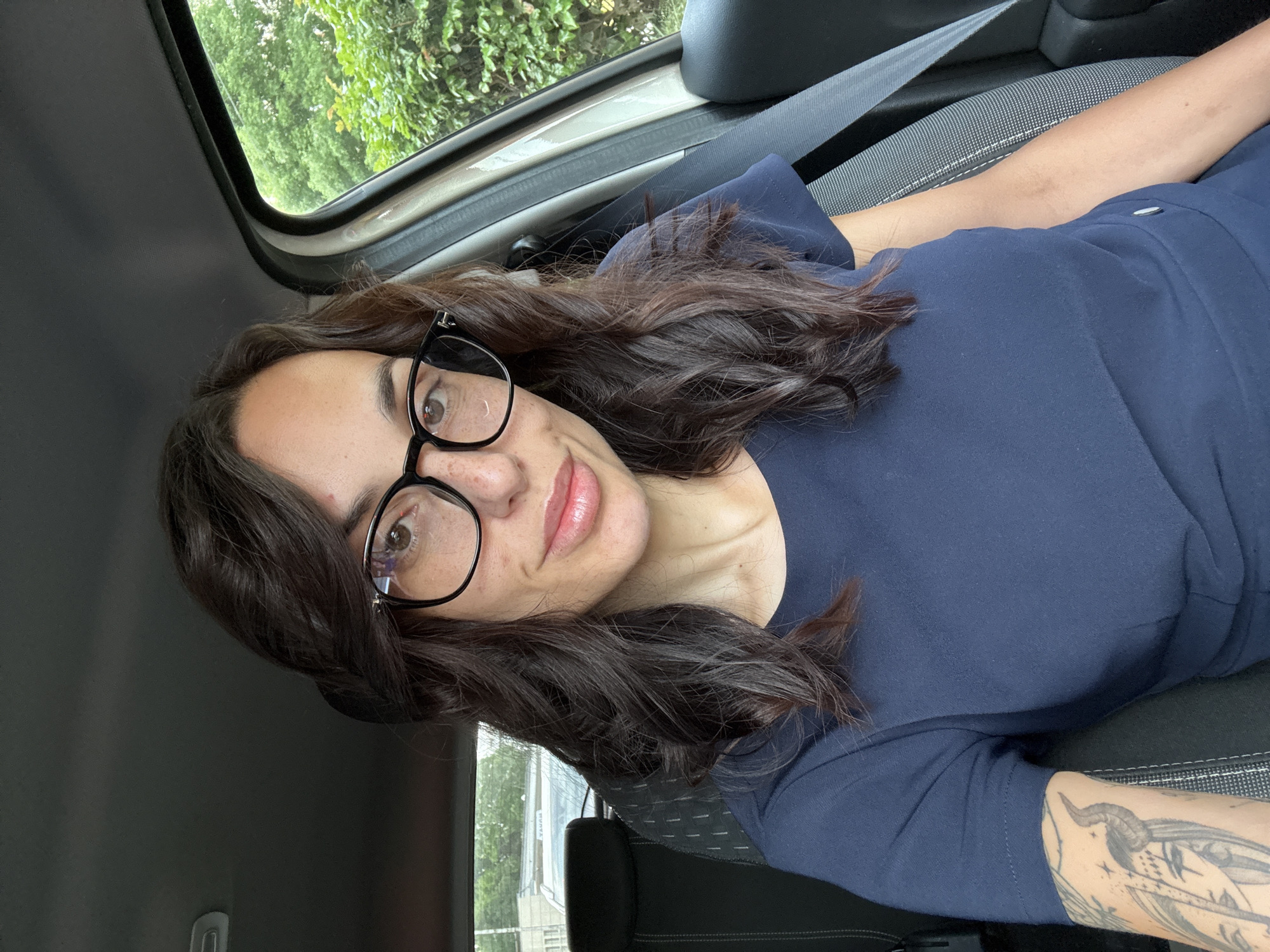 A woman is in a car. She is wearing glasses, a navy blue top, and a seatbelt. She has brown hair and fair skin. There is a tattoo on her forearm. The background shows a view through the car window, including trees and a sky that may be overcast. It is not possible to determine the specific location in Pleven, Bulgaria from this image.