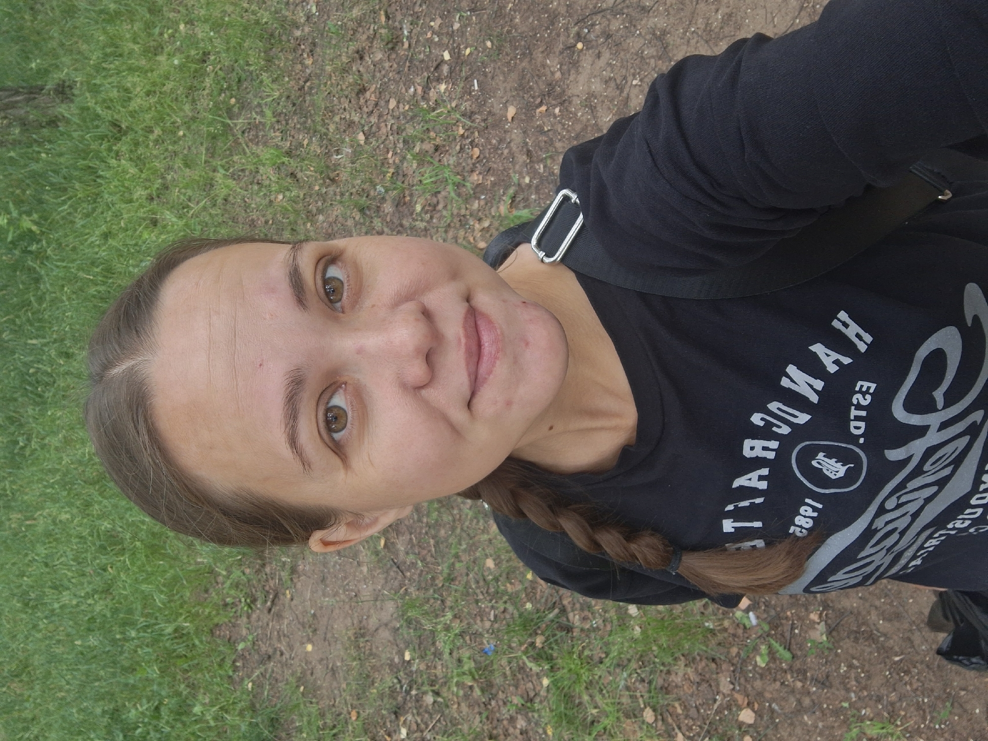 A young woman with brown hair braided down her left side and brown eyes is pictured from the chest up, looking directly at the camera. She is wearing a black t-shirt with a graphic design in a light color. The background consists of a dirt path with green grass on either side. The image appears to be taken outdoors in a park or natural area.