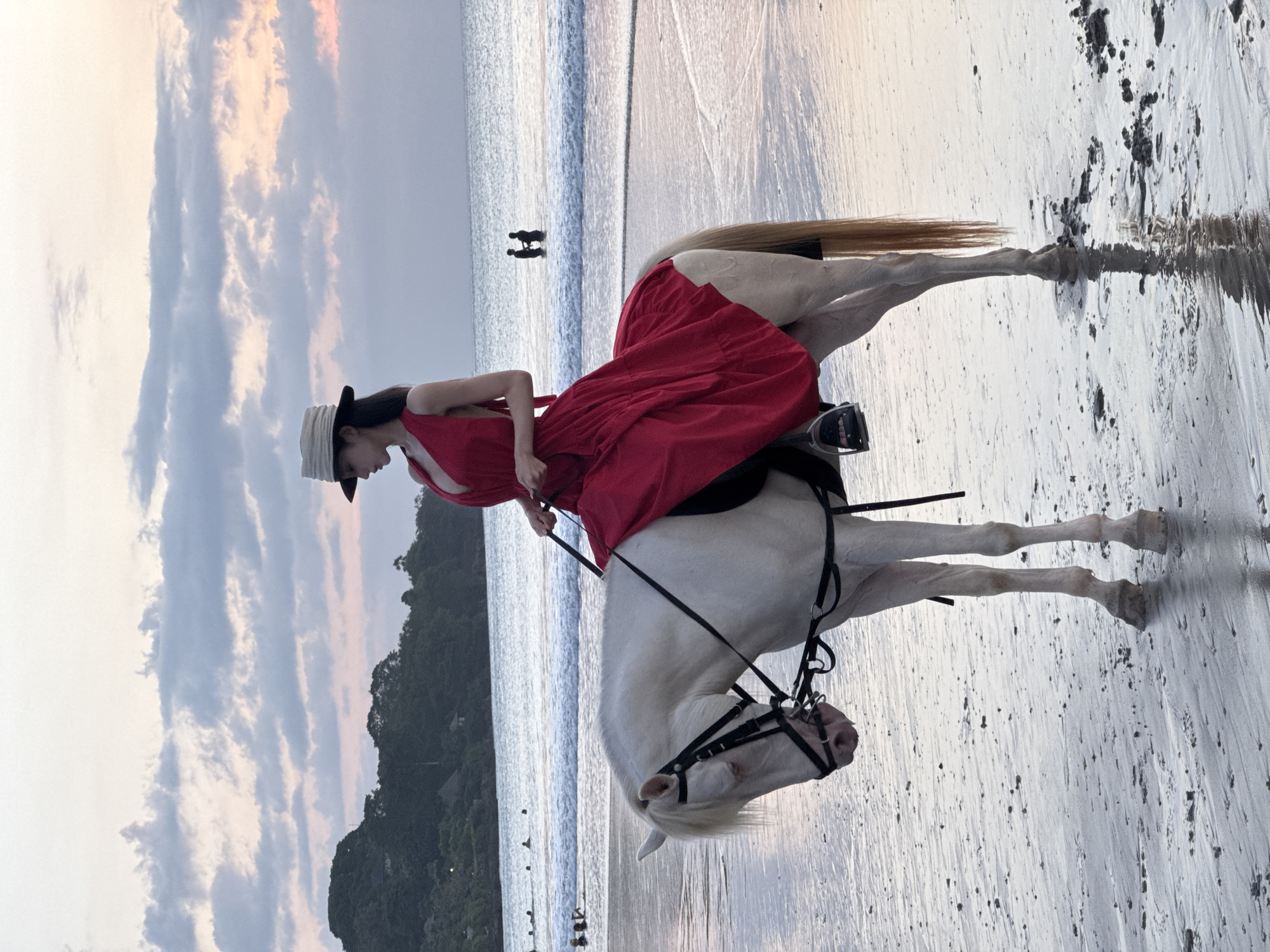 The image depicts a woman in a red dress and a straw hat riding a white horse on a wet, sandy beach. The horse is wearing a bridle. In the background, the ocean meets the shore, with gentle waves. Further out in the water, there are several people, some swimming and two standing in the water holding hands. In the distance, a tree-covered hillside forms the coastline, and the sky is filled with soft, pastel-colored clouds, suggesting either sunrise or sunset. The overall scene appears tranquil and picturesque, consistent with a beach location in Kuta Selatan, Indonesia, known for its scenic coastlines.