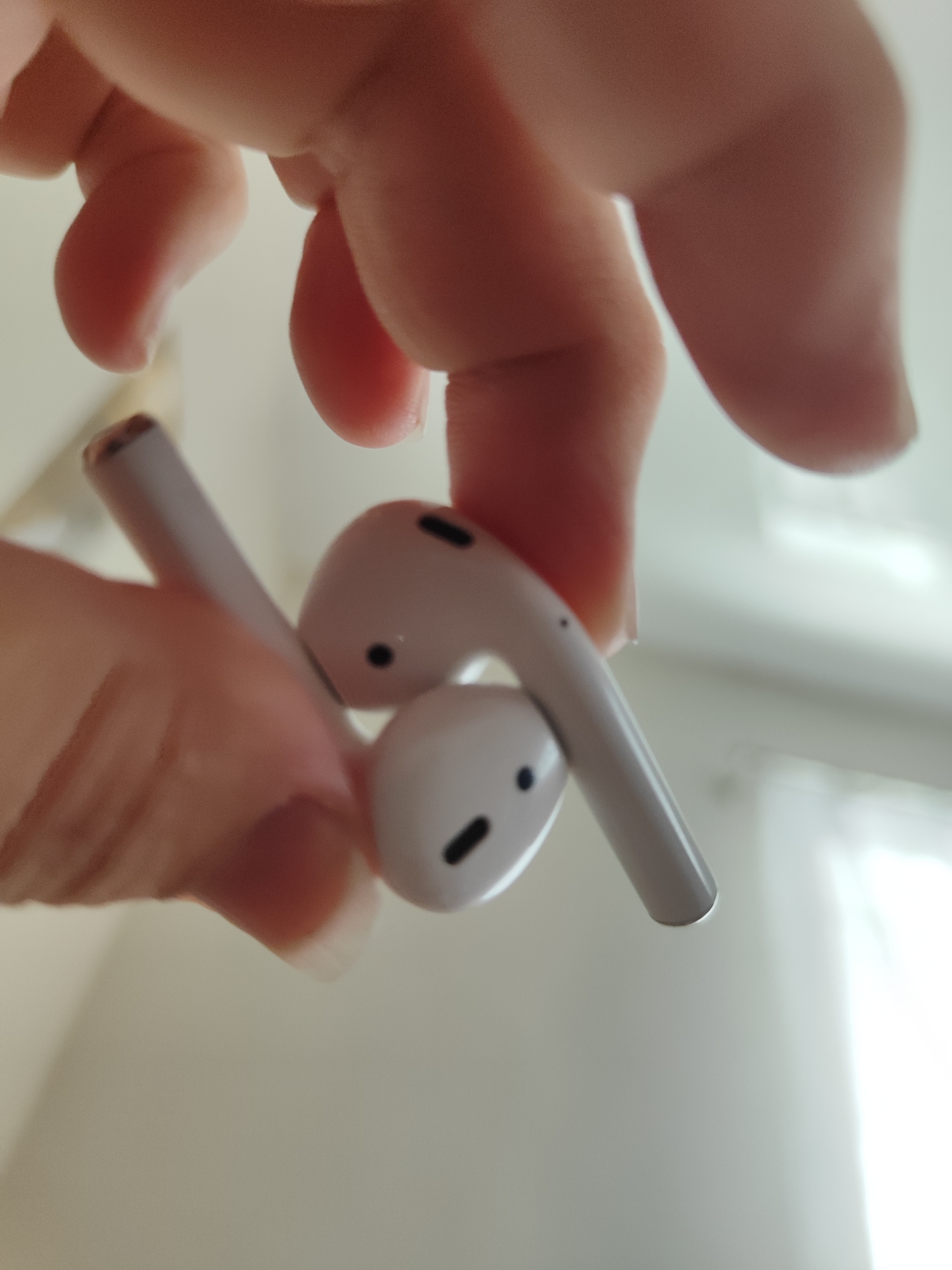 The image shows a close-up of a person's fingers holding two white wireless earbuds. The earbuds are the prominent objects, with one held in front of the other. The person's skin is visible, with hints of fingernails. The background is out of focus and appears to be an indoor setting with a bright, possibly artificial light source creating a diffused, almost white background. There are no visible text elements in the image. The focus is entirely on the earbuds and the fingers holding them, suggesting an activity related to handling or examining the earbuds. The lighting creates a soft, somewhat sterile atmosphere.