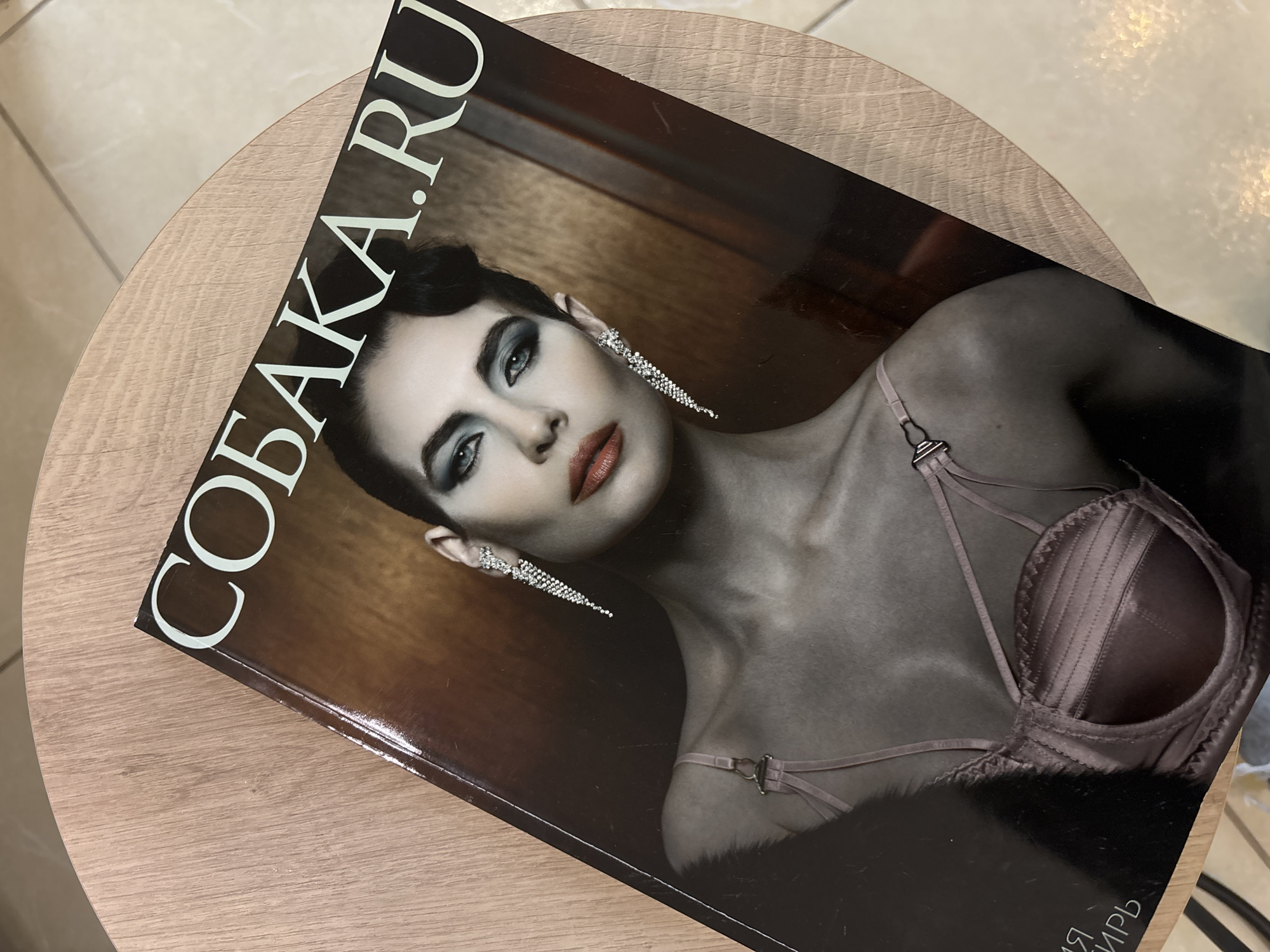 A close-up shot depicts a magazine cover featuring a woman with dark hair and striking makeup, including bold eyeshadow and red lipstick. She is wearing statement earrings and a bra-like garment, possibly lingerie. The magazine title "СОБАКА.RU" is prominently displayed. The magazine rests on a light-colored, round, wooden table. The location is Unknown, Russia.