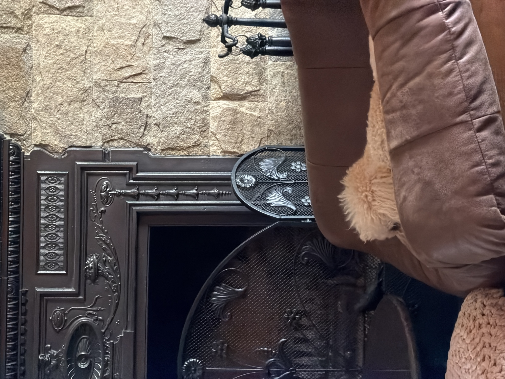 The image shows a stone wall partially obscured by a black, ornate fireplace with a curved grate. A brown cushion or furniture element is present on the right side, partially covering the fireplace. The image lacks context for location confirmation within Кирково, Bulgaria.