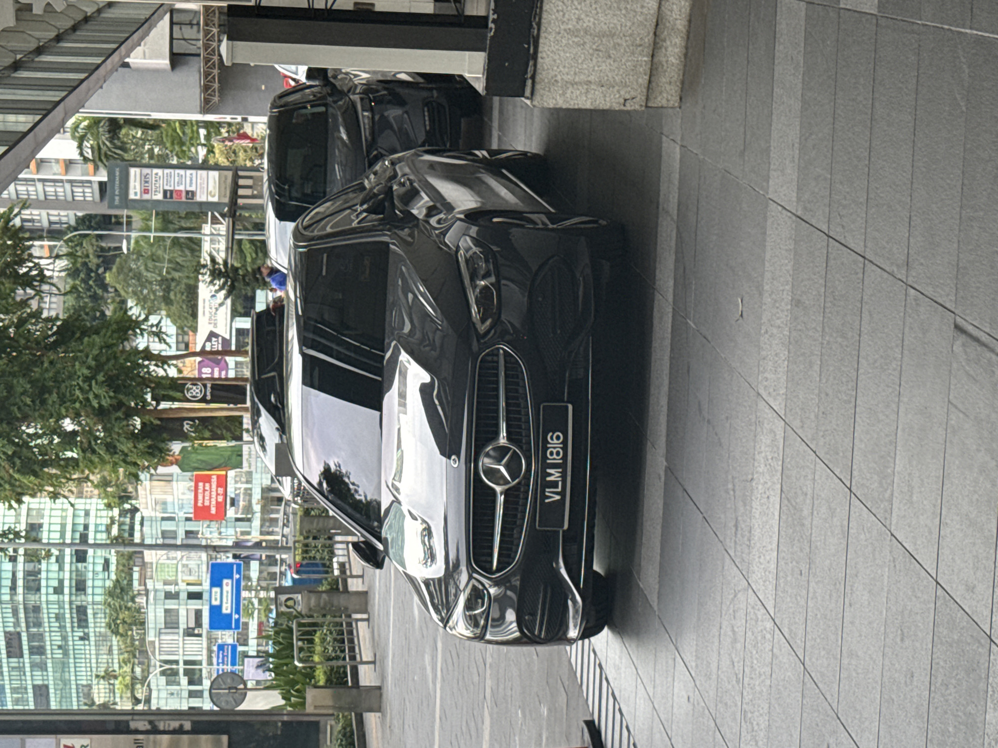 This daytime image showcases a black Mercedes-Benz SUV parked on a tiled pedestrian area next to a modern building in an urban setting. The car's front grille, headlights, and the Mercedes-Benz emblem are clearly visible, along with its Malaysian license plate reading "VLM 1816." Another dark-colored vehicle is partially visible behind it.
The background reveals tall, contemporary buildings with glass facades that reflect the surroundings, interspersed with green trees. A prominent street sign lists several establishments, including "THE INTERMARK," "ITRISS," "TSUTAYA," "DBS," "TOSCA," and "TONGA." Another red sign with white text is visible in the distance, partially reading "ANTARABANGSA 15-22," where "Antarabangsa" is the Malay word for "International," confirming the location as Kuala Lumpur, Malaysia. Additional visible text includes fragments like "EDUCATI LLEY DESTIMAT," "MERAN LAN," and "18 GPO." The ground is uniformly paved with grey rectangular tiles, and metal railings are present along the walkway. The scene suggests a vibrant commercial or mixed-use district in the city.
