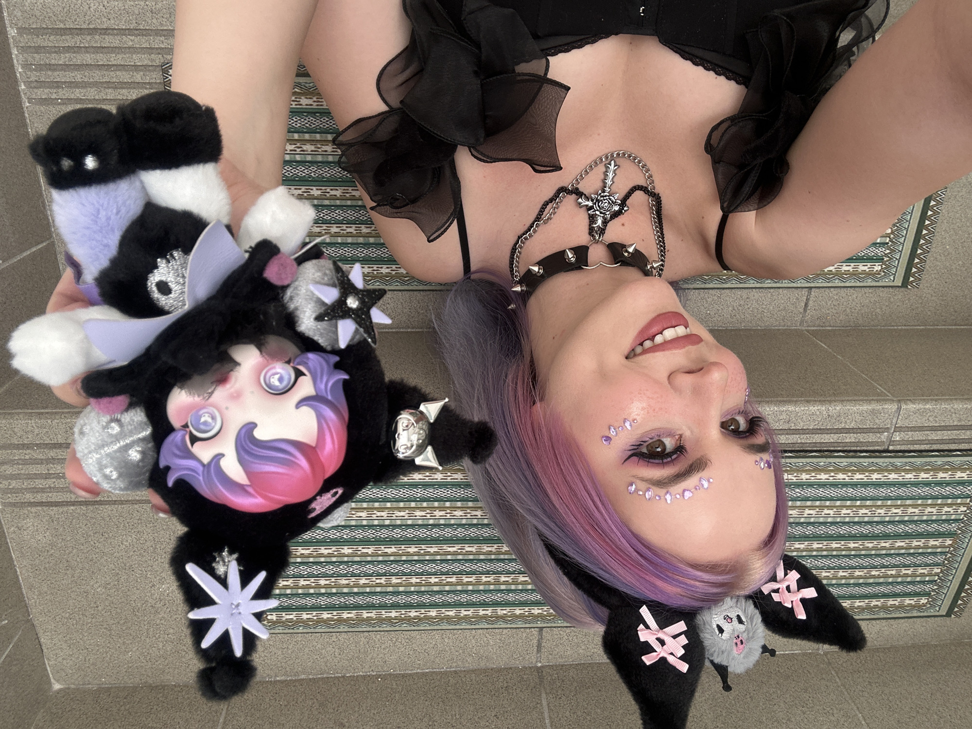 A person with pink and purple hair, wearing black cat ears and makeup with purple rhinestones, lies on a patterned mat on a gray tiled surface. They are holding a black and purple plush toy. The person is also wearing a black ruffled top, a black studded collar, and a chain with a rose cross. The image is taken from an upside-down perspective. The city mentioned in the request, City of Zagreb, Croatia, cannot be determined from the visual information provided in the image.