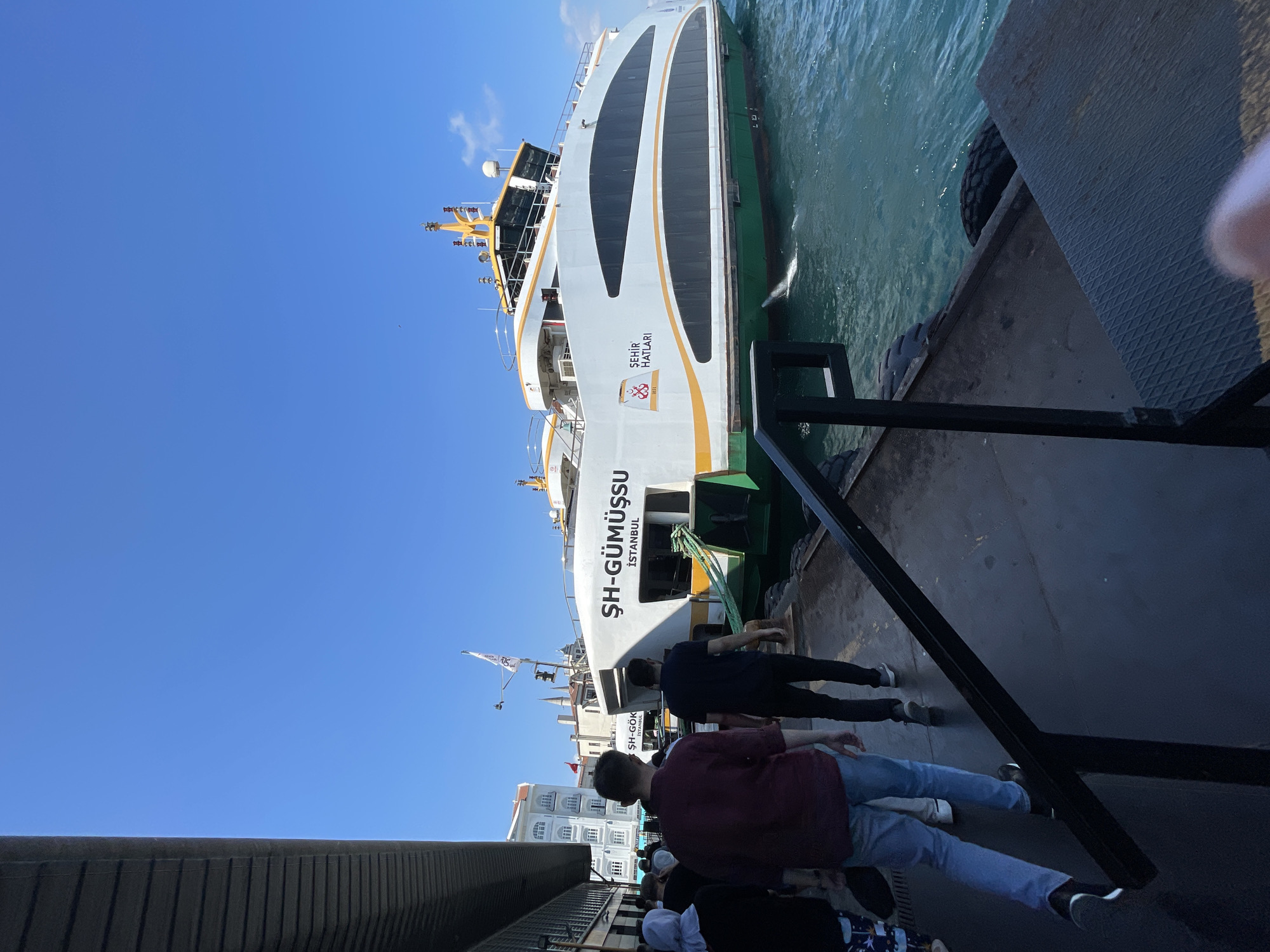 In Beyoğlu, Turkey, under a clear blue sky, a ferry named "ŞEHIR HATLARI" docks at a pier. The ferry, predominantly white with green and yellow accents, displays "ŞEHİR HATLARI" and "İSTANBUL" on its side. A green ramp extends from the ferry to the pier, suggesting boarding or disembarking. Several people, including a man in a maroon shirt and blue jeans, and another in a dark t-shirt and black pants, walk along the pier towards the ferry. A woman in a light-colored hijab is also visible among the group. The pier is made of dark material, with a black metal railing running alongside. The water around the ferry is a vibrant turquoise. Buildings can be seen in the background, including one with arched windows. The scene captures a moment of daily transportation in a bustling urban environment.