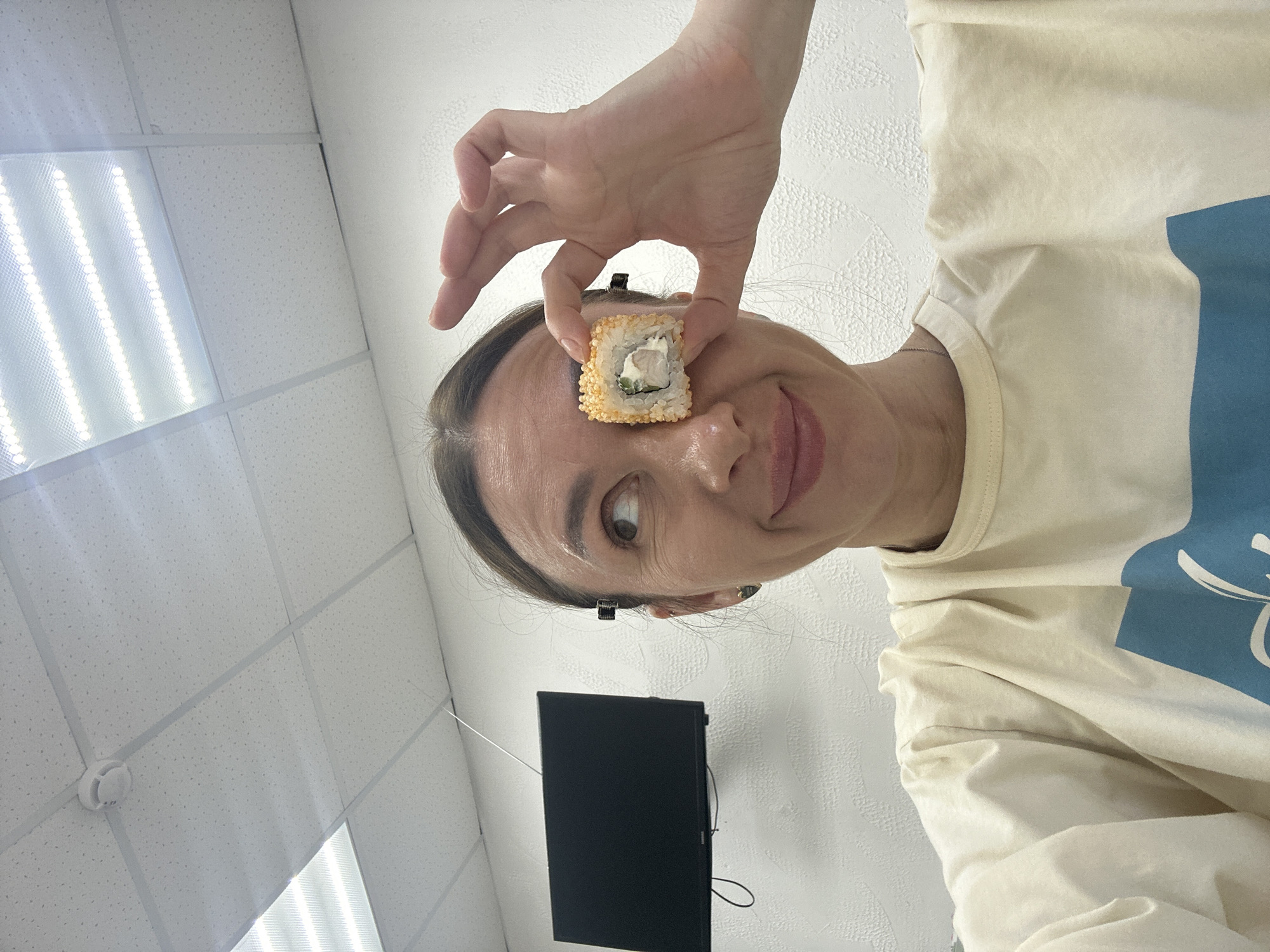A person is holding a piece of sushi in front of their forehead. The person is wearing a light-colored shirt with a blue design. The background includes a white tiled ceiling with fluorescent lights, a white wall, and a black rectangular object mounted on the wall (likely a television screen). It is not possible to determine whether this image was taken in Votkinsk, Russia.