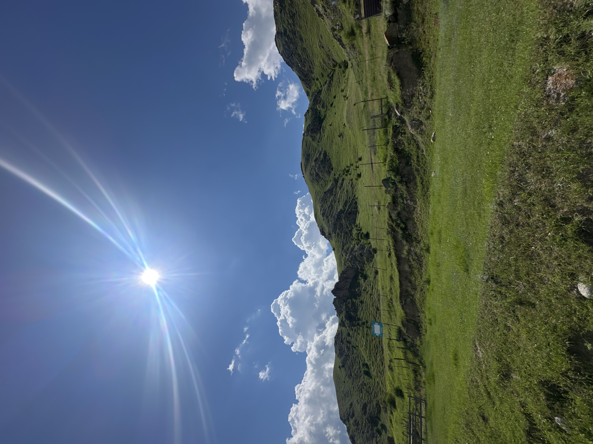 The image depicts a sunlit landscape in Bylym, Russia, characterized by rolling green hills under a bright blue sky with scattered white clouds. The sun is prominently visible in the upper left quadrant, casting strong rays and lens flare across the sky. The terrain is covered in lush green grass, with some rocky outcrops visible on the slopes. A wooden fence runs along the hillside, and a small, light blue sign is posted on the incline. The foreground features a grassy field with some rocks. The overall scene is one of natural beauty and openness.
