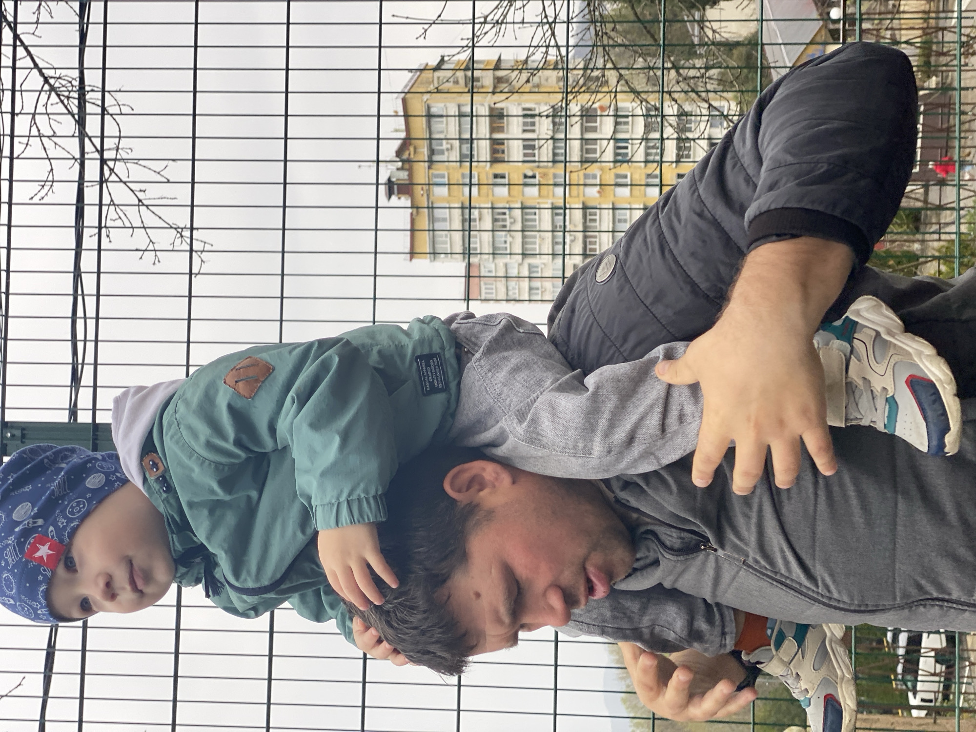 A man is carrying a young child on his shoulders in an outdoor setting. The child wears a blue hat with a red star, a light green jacket, and rests their hands on the man's head. The man wears a gray jacket and carries the child outside a metallic fence. In the background, an apartment building is visible, and branches of bare trees are seen through the fence. Given the architectural style of the apartment building and the common layout of residential areas, this is possibly located in Sochi, Russia.