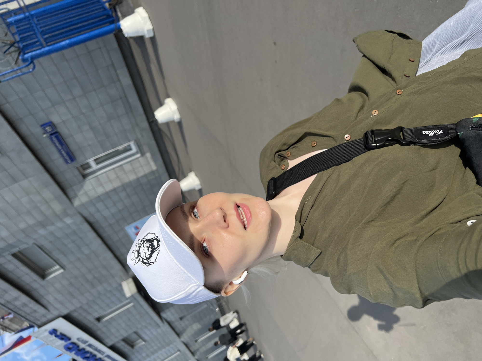 A woman stands on a city street in Khimki, Russia, angled to face the camera. She is wearing a white baseball cap with a black logo, an olive-green button-down shirt, and a black fanny pack. A light-blue striped garment is visible under the shirt. The street scene includes grey pavement, a blue metal structure, and a building with a street sign labeled in Russian. Several people are walking in the distance. The photograph appears to be taken in daylight, under sunny conditions.