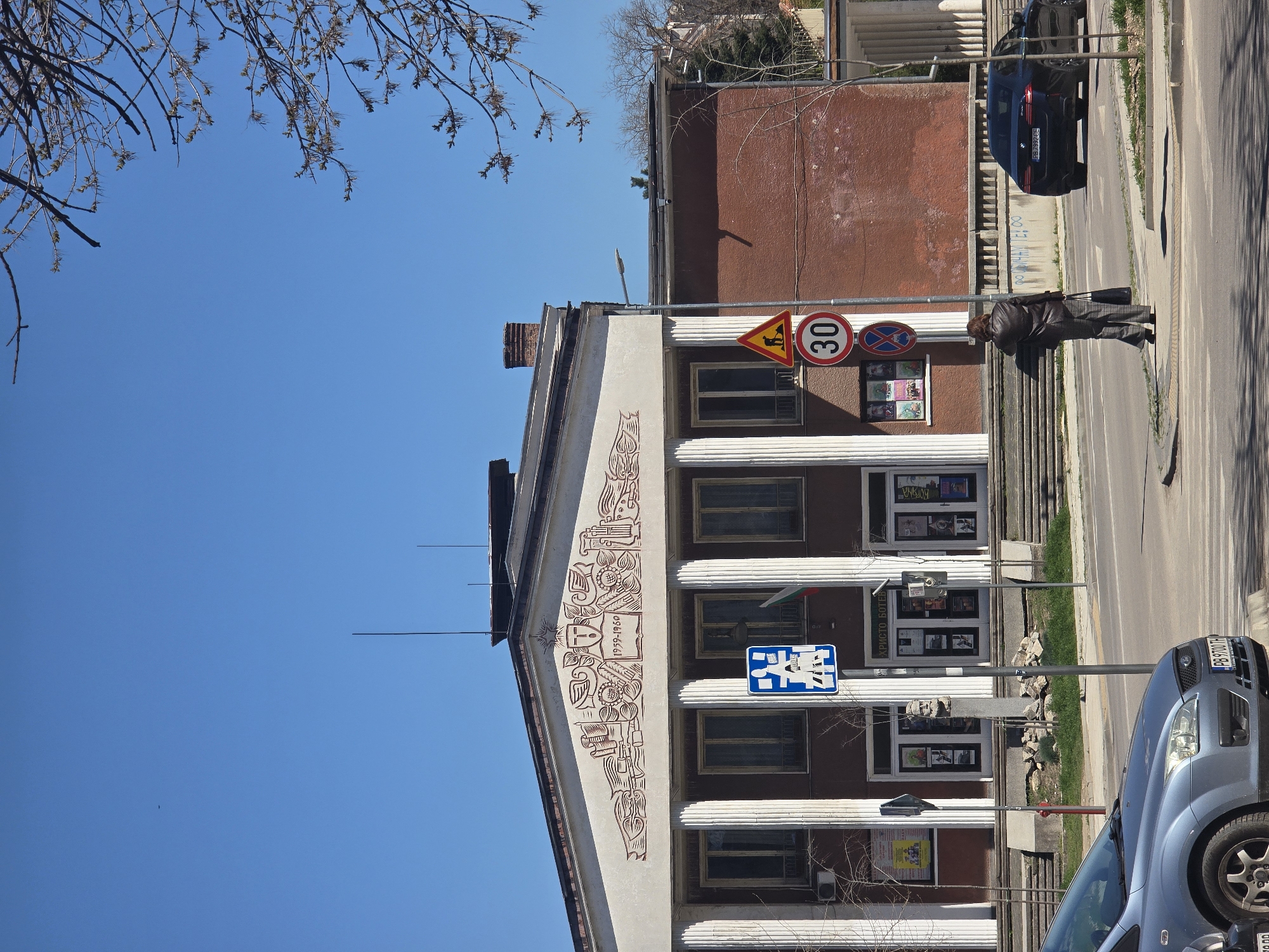 The image shows the exterior of the "Hristo Botev" community center building in Plovdiv, Bulgaria. The building features a facade with white columns and an embellished triangular pediment displaying a decorative mural dated 1959-1960. The building's facade is painted white, while the adjacent part of the building is brick-colored.
Several cars are parked on the street in front of the building. One is a blue car in the foreground with license plate PB 9700 TH, and another is a black car parked parallel to the curb. A pedestrian is walking on the sidewalk beside the building. Street signs are visible including a speed limit sign of 30 km/h.
The sky is bright blue and a tree branch extends into the upper left portion of the image.