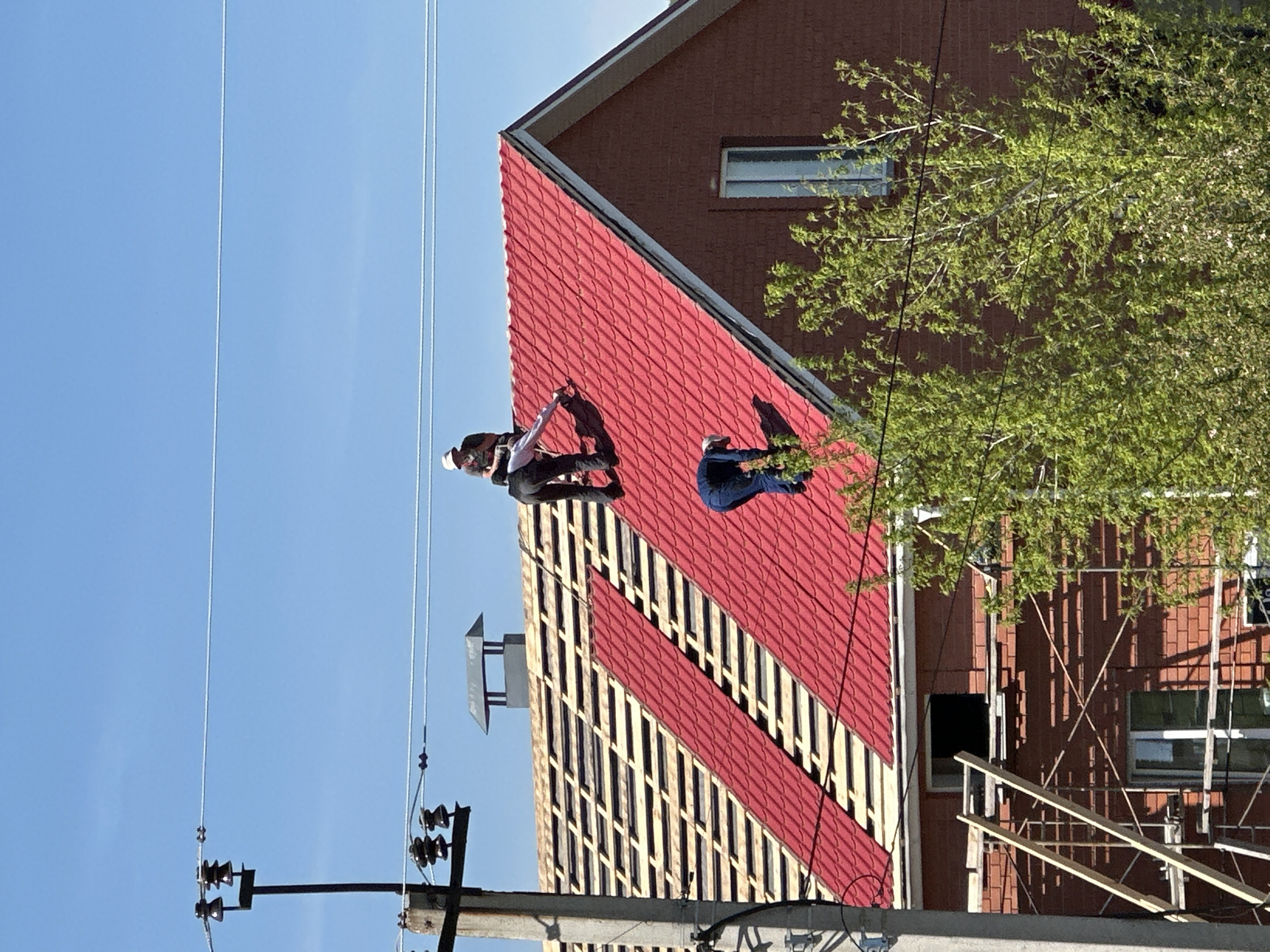A medium shot captures two people on a red-tiled roof, under a clear blue sky. The roof has a section with visible wooden beams underneath. A brick building forms the backdrop, with visible scaffolding and green foliage. Electrical poles and wires are in the foreground.
