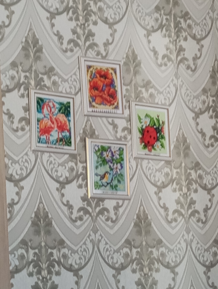 An interior wall in a residence located in the city of Яромаска, Russia, is depicted. The wall is covered with light-toned wallpaper featuring a repeating, ornate damask-style pattern in a slightly darker grey hue. Four square, light-colored framed artworks are affixed to the wall in a staggered, asymmetrical cluster. The artwork appears to be cross-stitch or diamond painting. The top-most frame displays red poppies with green leaves and blue accents. To the left and slightly below is a frame containing an image of two pink flamingos in a tropical setting. To the right of the poppy frame is a piece depicting a red ladybug on green foliage with small white flowers. Centered below the flamingo and ladybug frames is a fourth piece portraying a small brown and orange bird perched on a branch with purple flowers. No individuals, actions, or interactions are visible within the scene.