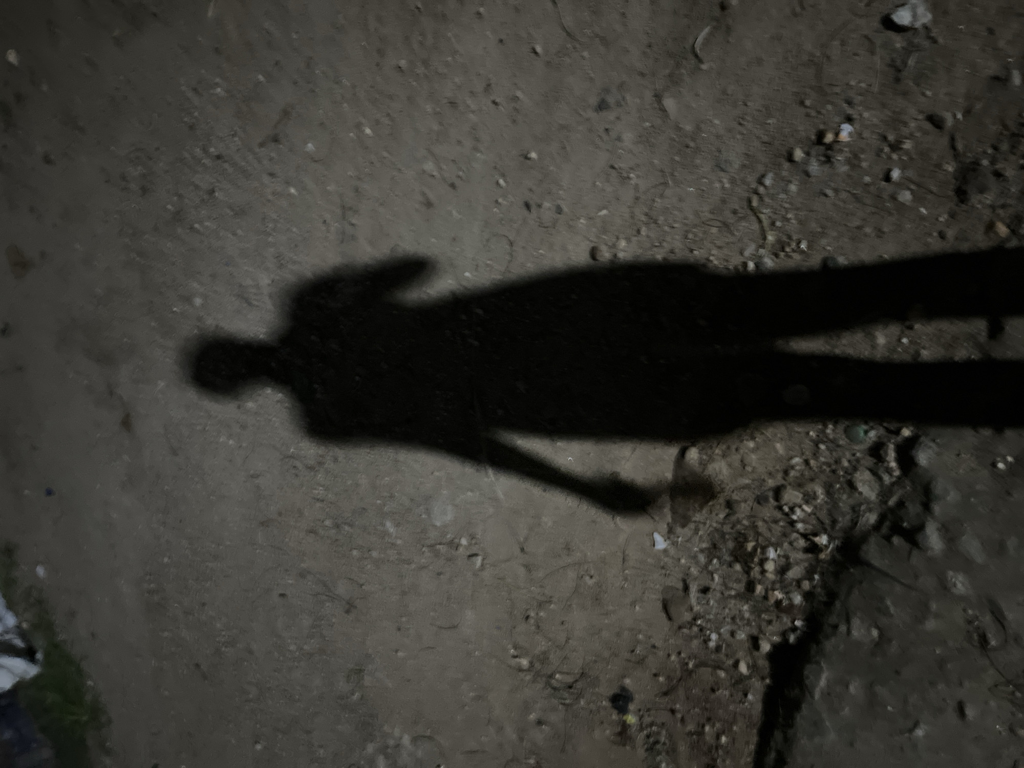A dark shadow of a person stands on a rough, uneven ground. The ground appears to be dirt or gravel, with small stones and debris scattered across its surface. In the background, there are hints of vegetation and possibly some discarded materials. The lighting suggests it is either night or a very dimly lit environment, casting a long, distinct shadow of the individual. The scene is devoid of any specific landmarks that would definitively place it in Abuja, Nigeria, but the texture of the ground and the general appearance are consistent with many outdoor urban or semi-urban areas within the city. There are no other people visible in the image, and the shadow suggests a solitary figure. The shadow itself shows the outline of a person, with a head, torso, arms, and legs. One arm appears to be slightly raised. The environment is dark and somewhat unkempt.