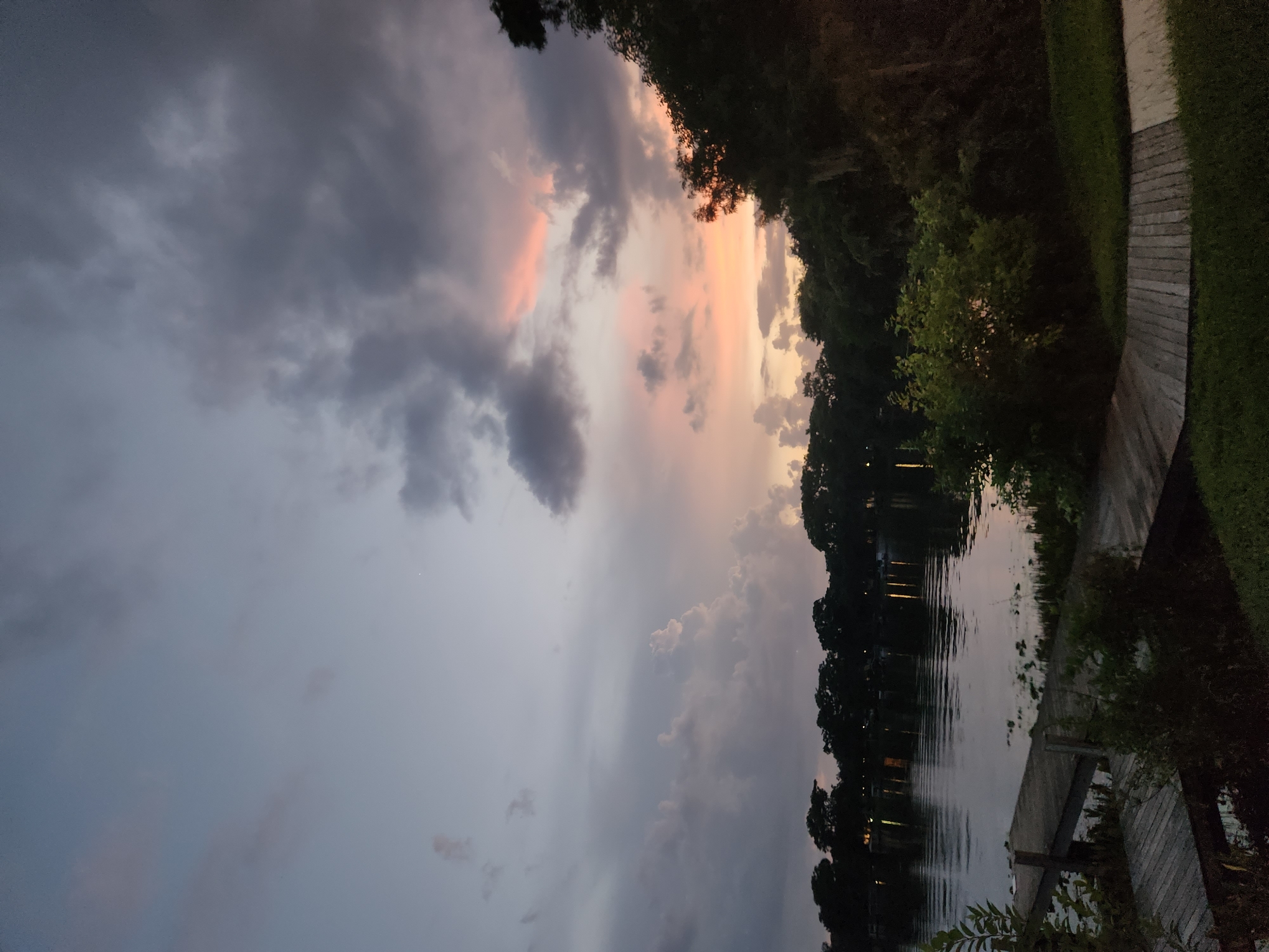 The image depicts a serene twilight scene at Lake Osceola in Winter Park, Florida, USA. A wooden dock extends into the calm water, which reflects the dusky sky and the silhouettes of trees lining the opposite shore. The sky is a canvas of muted blues and grays, interspersed with soft pink and orange hues from the setting sun. Wispy clouds drift across the horizon, adding depth and texture to the atmospheric portrayal of the end of the day. The foreground features lush green grass and dense foliage, with parts of the wooden dock also visible. No individuals are present in the scene.