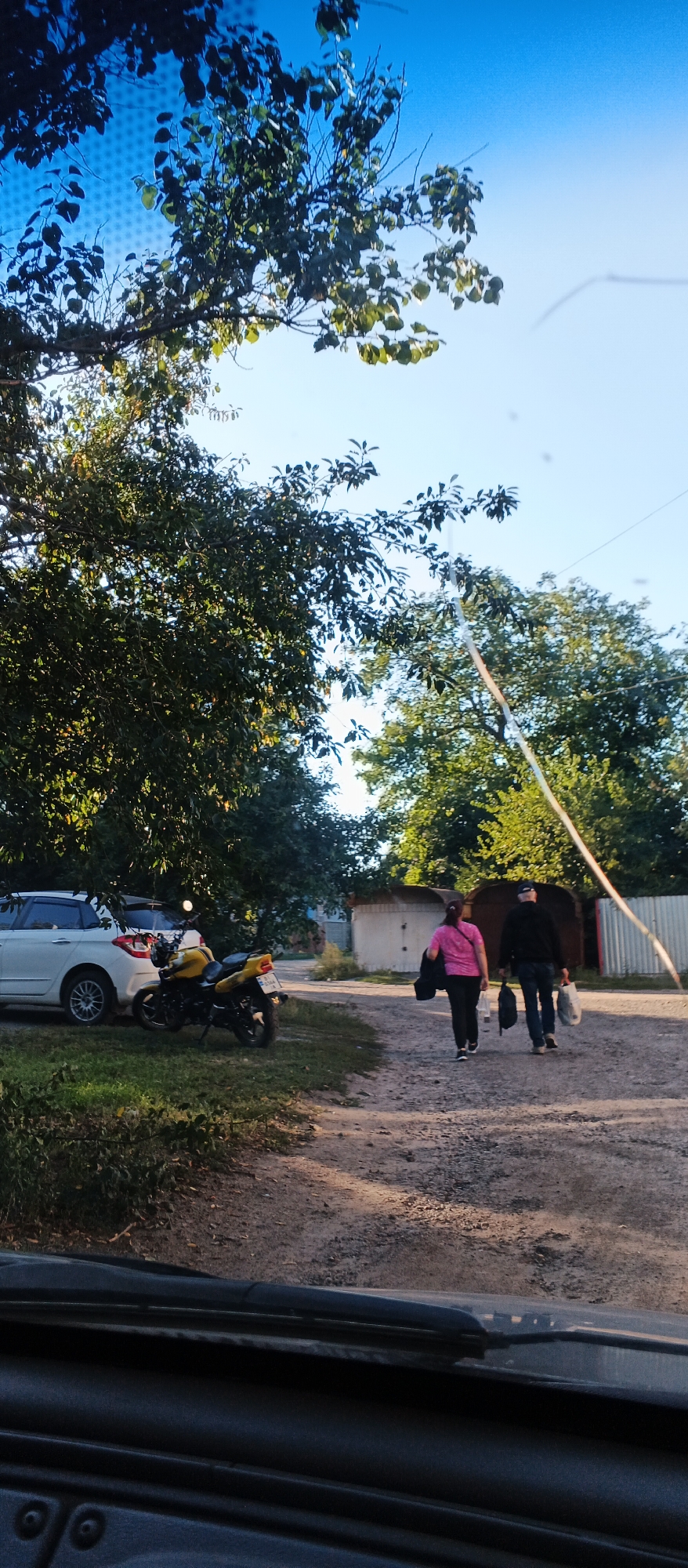 In Smila, Ukraine, a scene unfolds on a dirt road under a clear blue sky, bordered by trees. On the left, a white car is parked next to a vibrant yellow motorcycle. Two individuals, a woman in a pink shirt and a man in a dark jacket and jeans, walk away from the camera towards the right of the frame, each carrying bags. In the background, buildings and a corrugated metal fence line the area, suggesting a residential or semi-industrial setting. The ground is covered with loose gravel and dirt, with patches of grass and greenery visible. The perspective suggests the photo was taken from inside a vehicle, possibly looking through a windshield.