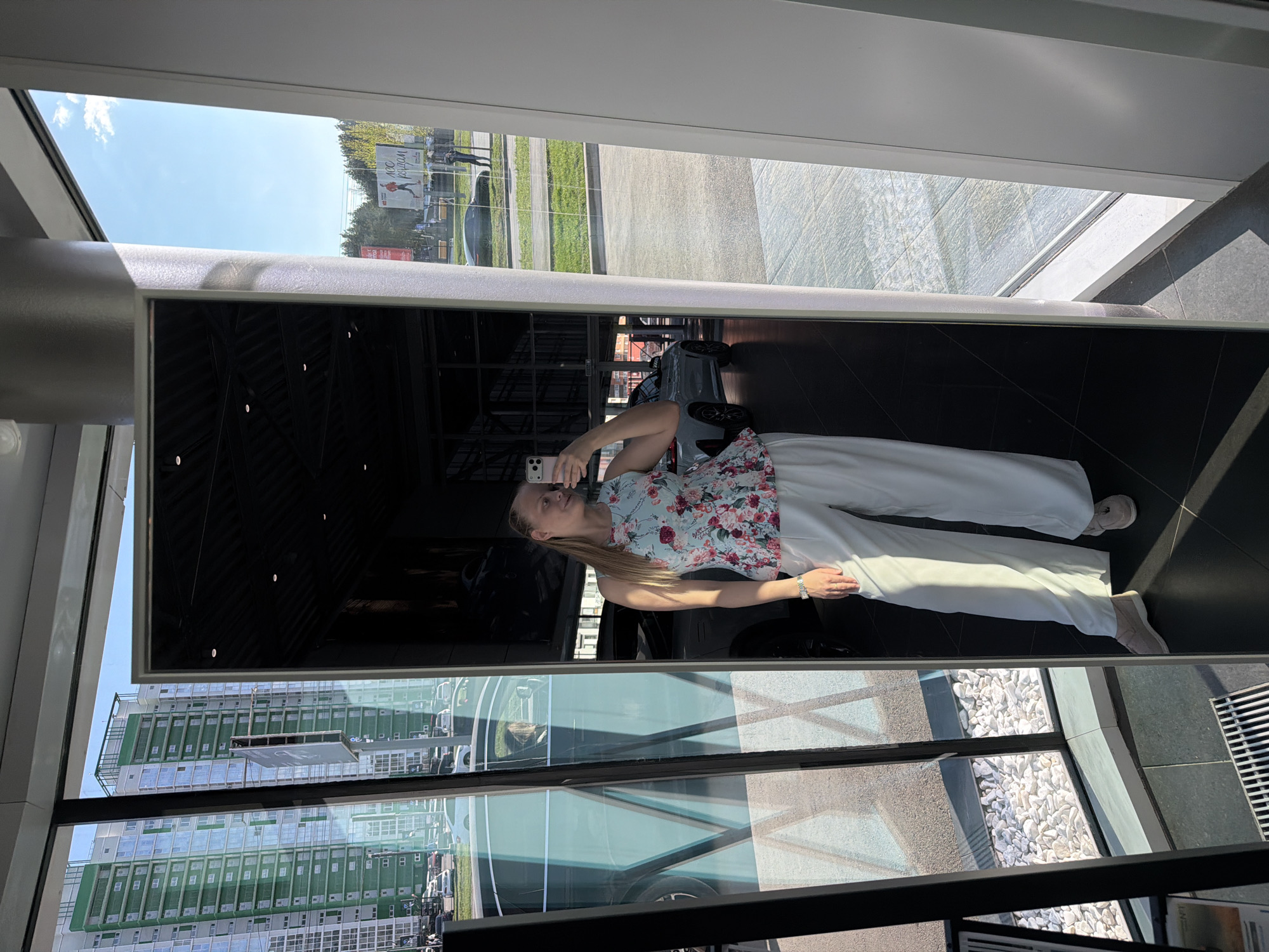 A woman is taking a selfie in a mirror. She is wearing a floral print top and white pants. The mirror reflects a modern building with large windows, a view of a city, and a glimpse of the sky. The reflection includes a partial view of a car and some landscaping elements. The location is Izhevsk, Russia, based on visible architectural features and city planning.
