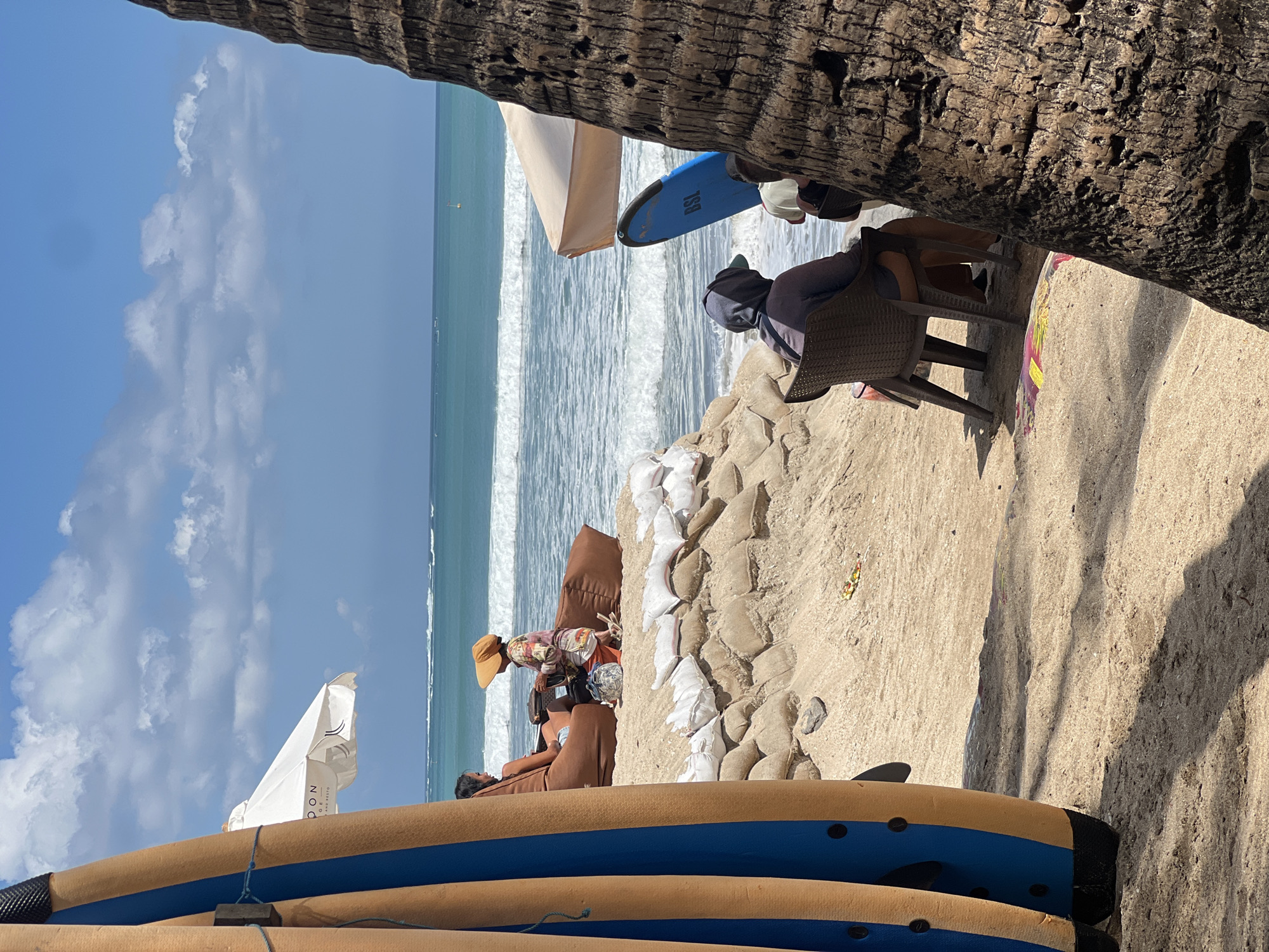 This is a wide shot of a beach in Kuta, Indonesia, on a bright, sunny day with scattered clouds. In the foreground, a large palm tree trunk is on the right, obscuring part of the view. To the left, two surfboards are propped up. A sandbag wall runs horizontally across the middle ground, creating a raised platform.
On top of the sandbag wall, two people are seated on large, brown beanbag chairs. One person, wearing a wide-brimmed hat, is facing away from the camera, looking towards the ocean. The other person is partially visible, also looking out at the water.
Further back, on the sandy beach, two more individuals are seated in plastic chairs, facing the ocean. One person is wearing a hooded garment. A blue surfboard with "BSL" written on it stands upright behind them.
The ocean in the background features moderate waves and a clear blue sky with white clouds. A few distant boats or buoys are visible on the water. The overall scene depicts a relaxed beach atmosphere with elements of surfing and leisure.