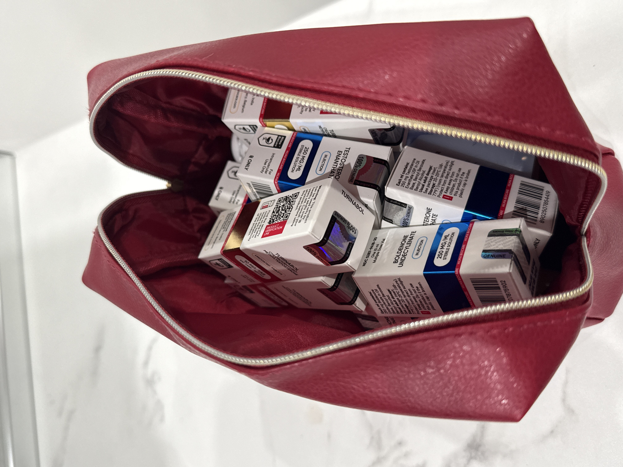 An open, red, textured zippered bag, possibly made of faux leather, is shown filled with numerous small, uniformly packaged boxes. The bag rests on a light-colored surface against a white background. The contents are pharmaceutical or supplement products, primarily identifiable as anabolic steroids from the brand "HILMA BIOCARE."
Visible text on the boxes identifies several products: "TESTOSTERONE ENANTHATE" (250 MG/ML STERILE SOLUTION for INJECTION), "BOLDENONE UNDECYLENATE" (250 MG/ML STERILE SOLUTION for INJECTION), and "TURINABOL" (CHLORMETHYLTESTOSTERONE 10 MG/TAB UNCOATED TABLETS). The boxes also display dosage and usage instructions, such as "Each ml contains: 250 mg Testosterone Enanthate USP in Oil," "Usual adult dosage: See package insert," and storage conditions like "20-25°C." Warnings are present, including "Keep away from children" and "Do not use this product if the plastic cap is broken." Some labels also state "For Intramuscular Use Only" and "R ONLY." Repeated declarations of "100% GENUINE" and "PRODUCT VERIFICATION LINKs" with QR codes suggest measures for authenticity. Other visible text includes "NDC 0287-9418-30," "8902507854419," "07875902," and mentions of "India" and "Biocare.com." The scene is a static display of the bag's contents. While the location context of Burgas, Bulgaria is provided, no visual cues in the image corroborate this.