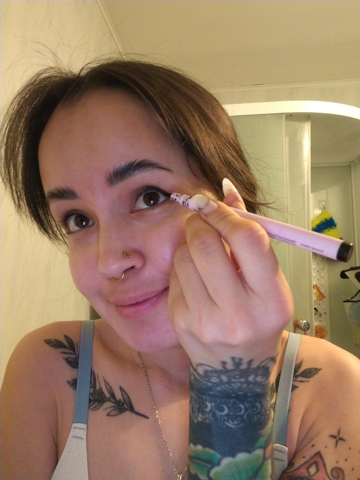 A selfie of a woman with brown hair wearing a light blue bra and a silver necklace, applying eyeliner to her right eye. She has a nose ring on the left side of her nose and two additional piercings on the left side of her nose. She has multiple tattoos on her left shoulder and arm. The background appears to be a bathroom, with a white shower curtain and a yellow and blue sponge hanging on a hook. The location is unknown.