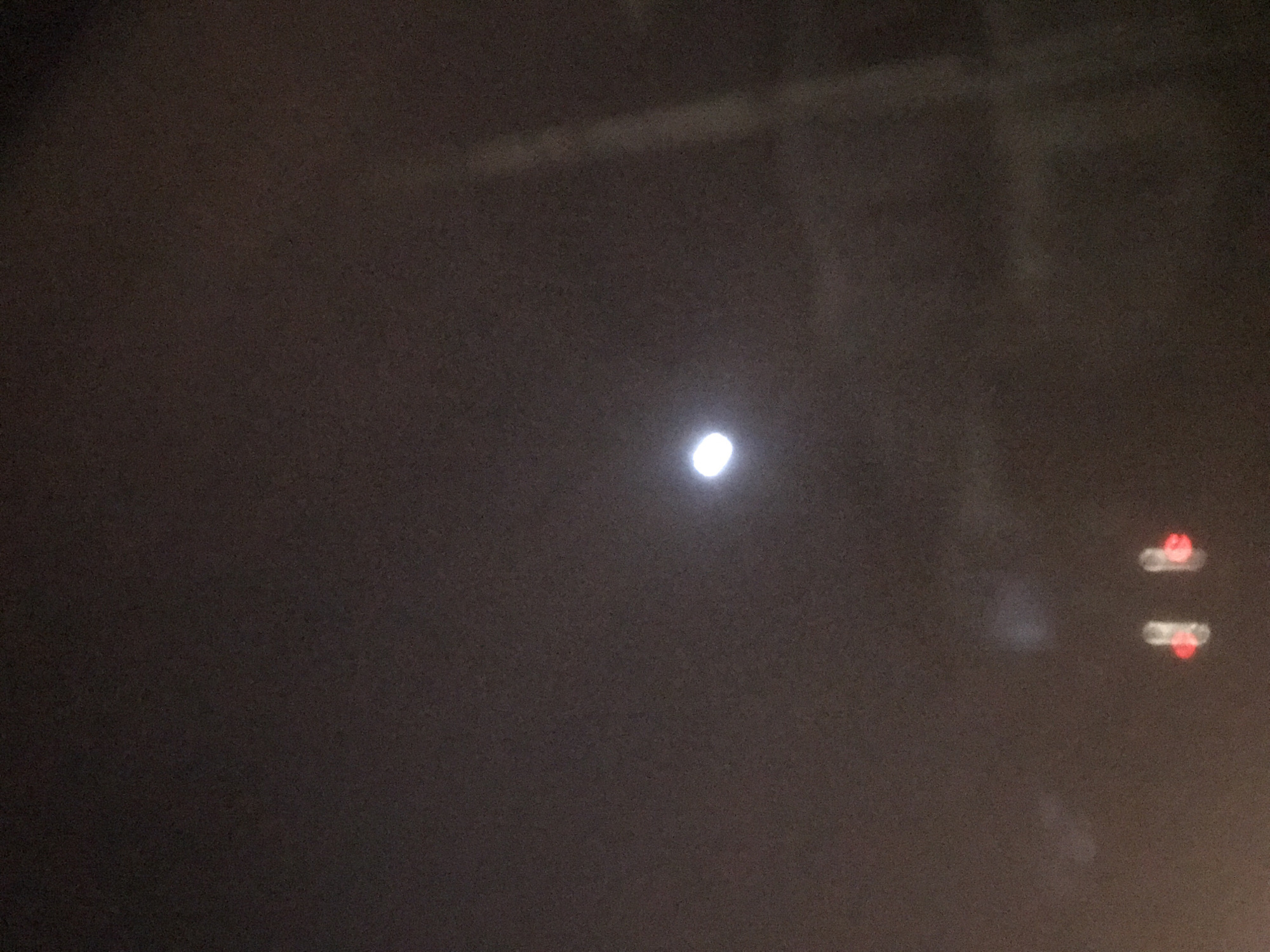 The image depicts a dark, hazy scene with a bright, circular light source near the center. To the right of this central light, there are two distinct blurry lights, one above the other, appearing to be red and white. The overall lighting is dim, suggesting it is nighttime or a very dark environment. The haziness could indicate fog, smoke, or simply a low-quality image capture. There are no discernible people, animals, or specific objects besides the lights. The background appears to be a uniform, dark color, possibly the sky or a darkened interior. The bright central light might be the moon or a distant streetlamp, while the lights on the right could be from vehicles or another light source. The image conveys a sense of mystery due to the lack of clear details and the indistinct nature of the light sources.