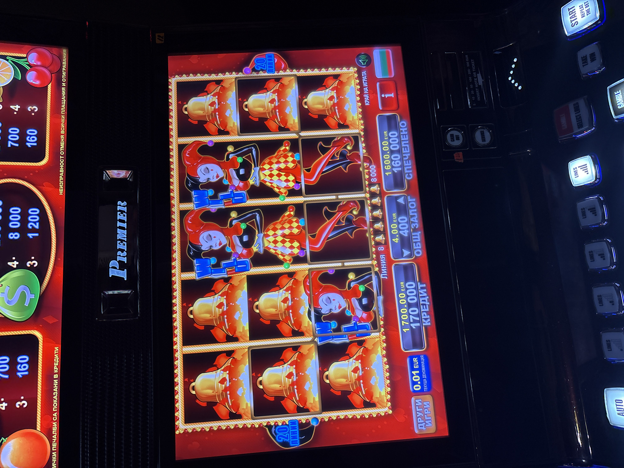 The image shows a close-up, slightly angled interior shot of a slot machine within a casino or gaming establishment. The slot machine's screen dominates the frame, displaying a vibrant red interface with various game symbols, including bells and jester-like characters. Numerical displays indicating credits, bet amounts, and winnings are visible in EUR. The machine is active, with illuminated buttons on the control panel below the screen, including "AUTO," "LINES," "SELECT LINES," "GAMBLE," "TAKE WIN," and "START." The overall environment appears dark, typical of a casino, with some ambient light reflecting off the machine's surfaces. The machine is labeled "PREMIER" at the top. The context suggests a scene within a casino in Sopot, Bulgaria, likely a modern gaming establishment. There are no people clearly visible in the frame, but the presence of an active slot machine implies the potential for patrons to be nearby. The machine itself appears to be in good working order.