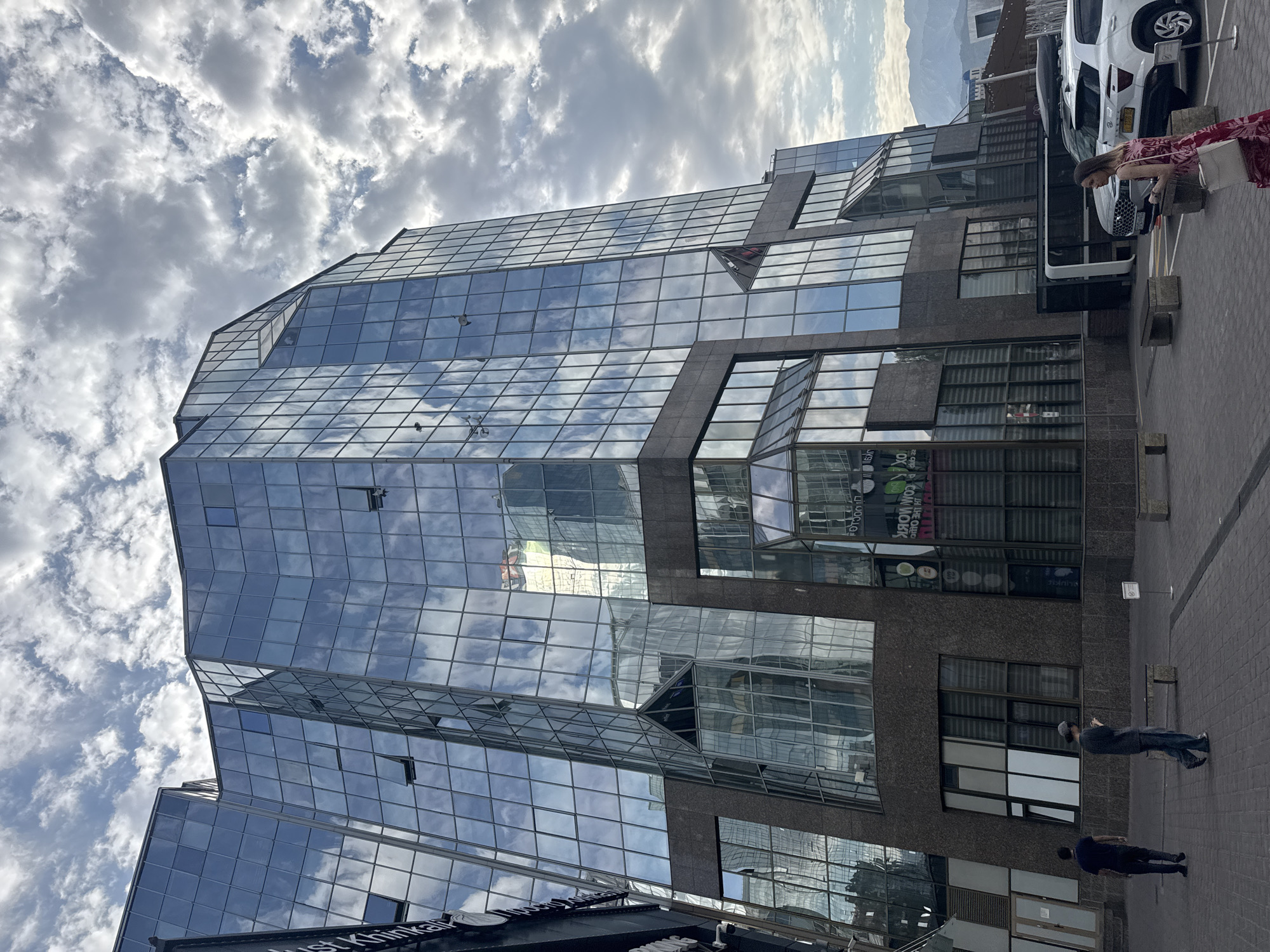 A low-angle, exterior, medium shot captures a modern, multi-story glass building with a unique, angular design. The building's facade is composed of numerous reflective glass panels, creating a mirrored effect of the cloudy sky and surrounding architecture. Several windows are slightly ajar. The lower floors of the building feature a darker, stone-like material. On the ground level, a paved walkway leads past the building. Two men are walking away from the camera on the left side of the walkway. Further right, a woman in a patterned dress is walking towards the camera, carrying a bag. Parked cars, including a white SUV and a grey vehicle, are visible behind her. In the background, mountains are visible against the horizon under a cloudy sky. The scene appears to be in the city of Almaty, Kazakhstan, likely in a commercial or business district.