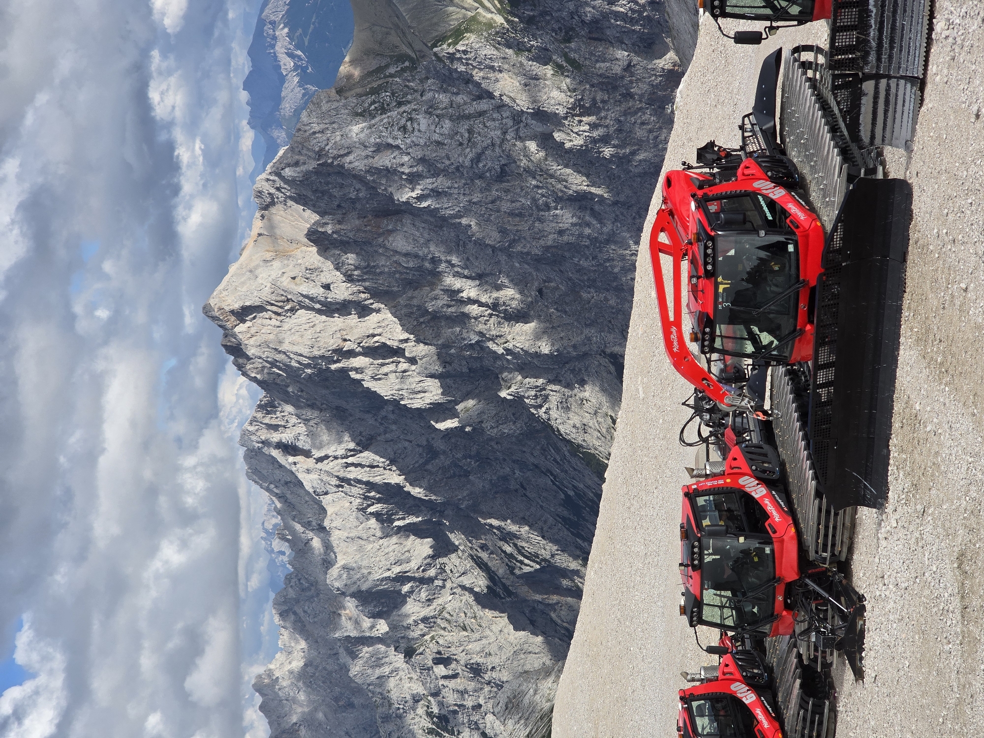 The image depicts a line of red snow groomers parked on a gravelly surface with a majestic mountain range in the background. The snow groomers, specifically Prinoth brand models, feature large tracks and blades, indicative of their purpose in maintaining ski slopes. One groomer is more prominently featured in the foreground, showcasing its bright red exterior and the "640" model designation. The mountain landscape is characterized by rugged, gray rock faces and distant peaks under a cloudy sky with patches of blue. The scene is likely located at a ski resort or a high-altitude area near Garmisch-Partenkirchen, Germany, given the presence of specialized snow maintenance equipment and the alpine terrain. There are no people visible in the frame.