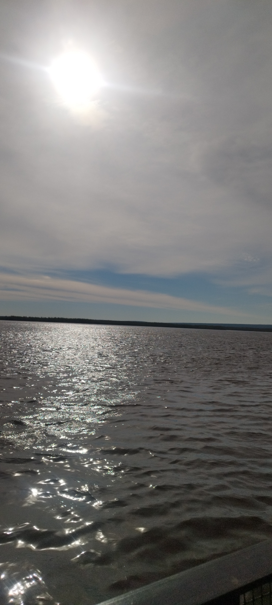 The image shows a body of water under a sky with a bright sun obscured by clouds. The water surface reflects sunlight, creating shimmering patterns. The horizon line is visible with a dark strip of land in the distance. The location cannot be determined from the image. No people, interactions or other objects are visible.