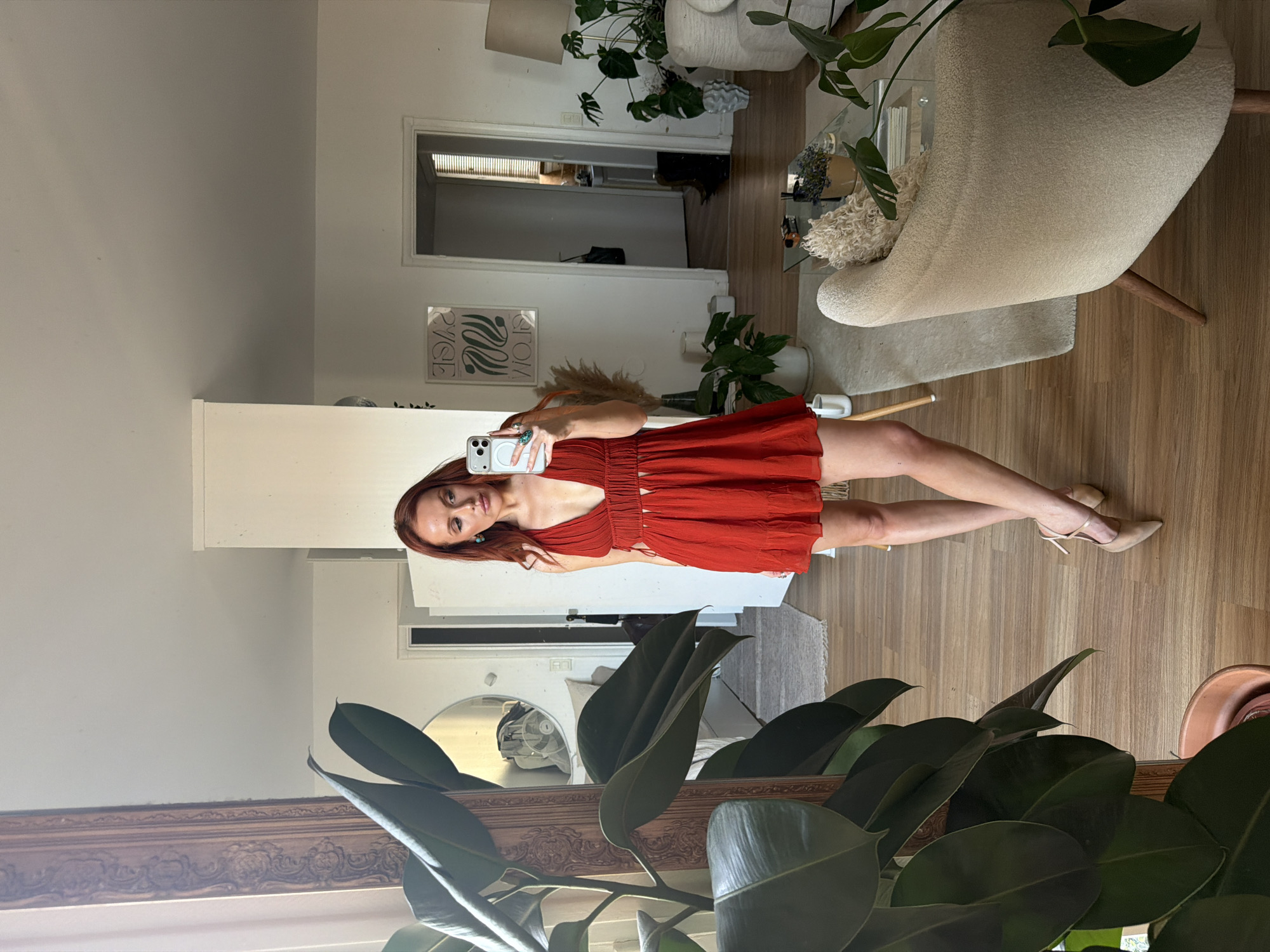 A fair-skinned woman with red hair is standing in a well-lit room, taking a mirror selfie. She is wearing a short, red dress and beige heels. Her phone is visible in her right hand, and she is looking at the screen.
The room is decorated with plants, a beige chair, and a glass coffee table. The floor is made of wood. A doorway is visible in the background, leading to another room. The mirror is framed in an ornate, wooden frame. There is no evidence to suggest this photo was taken in Paris, France.