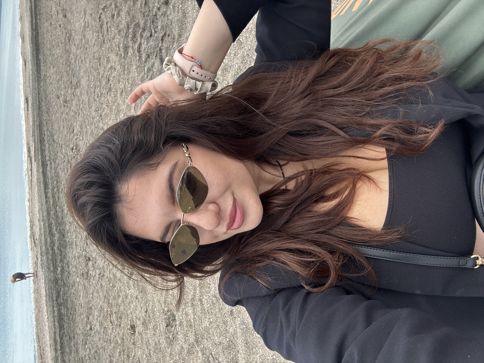 A woman with long brown hair, wearing sunglasses and a black shirt, is lying on a sandy beach. She has a watch and scrunchie on her left wrist. In the background, there is a person standing on the beach near the water. The location is Burgas, Bulgaria, as indicated by the dark sand.