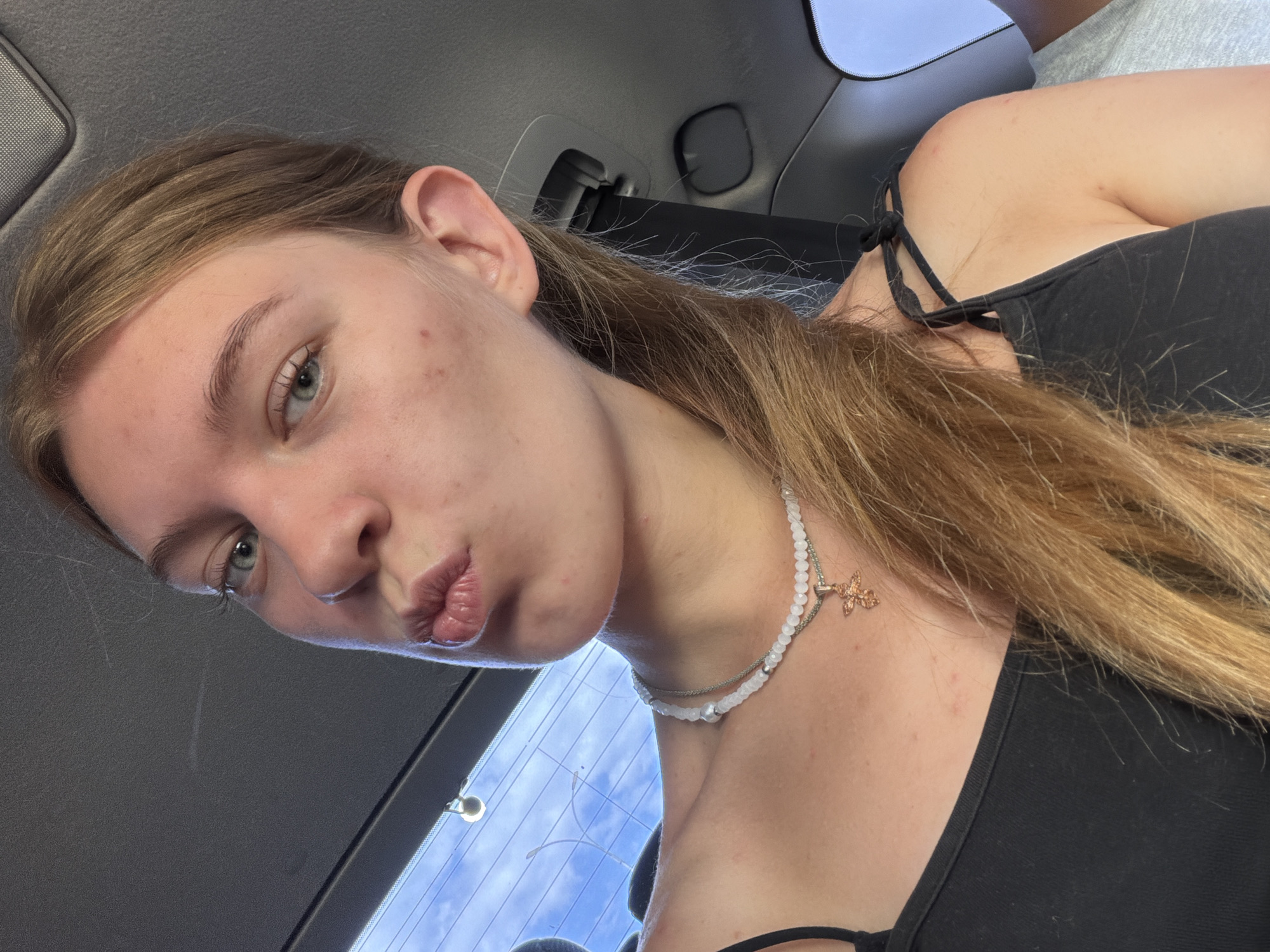 A young woman with blonde hair is captured in a selfie taken from the shoulders up. She is wearing a black tank top and a beaded necklace with a cross pendant. Her lips are pursed in a "duck face" pose, and her eyes are looking towards the camera. The background suggests she is inside a vehicle, with the interior of the car visible, including a seatbelt buckle and a glimpse of the sky and trees through a window. The lighting indicates it is daytime. The image is a close-up shot focusing on the woman's face and upper body. There are no identifiable landmarks or contextual clues to suggest the location is Rostov-on-Don, Russia.