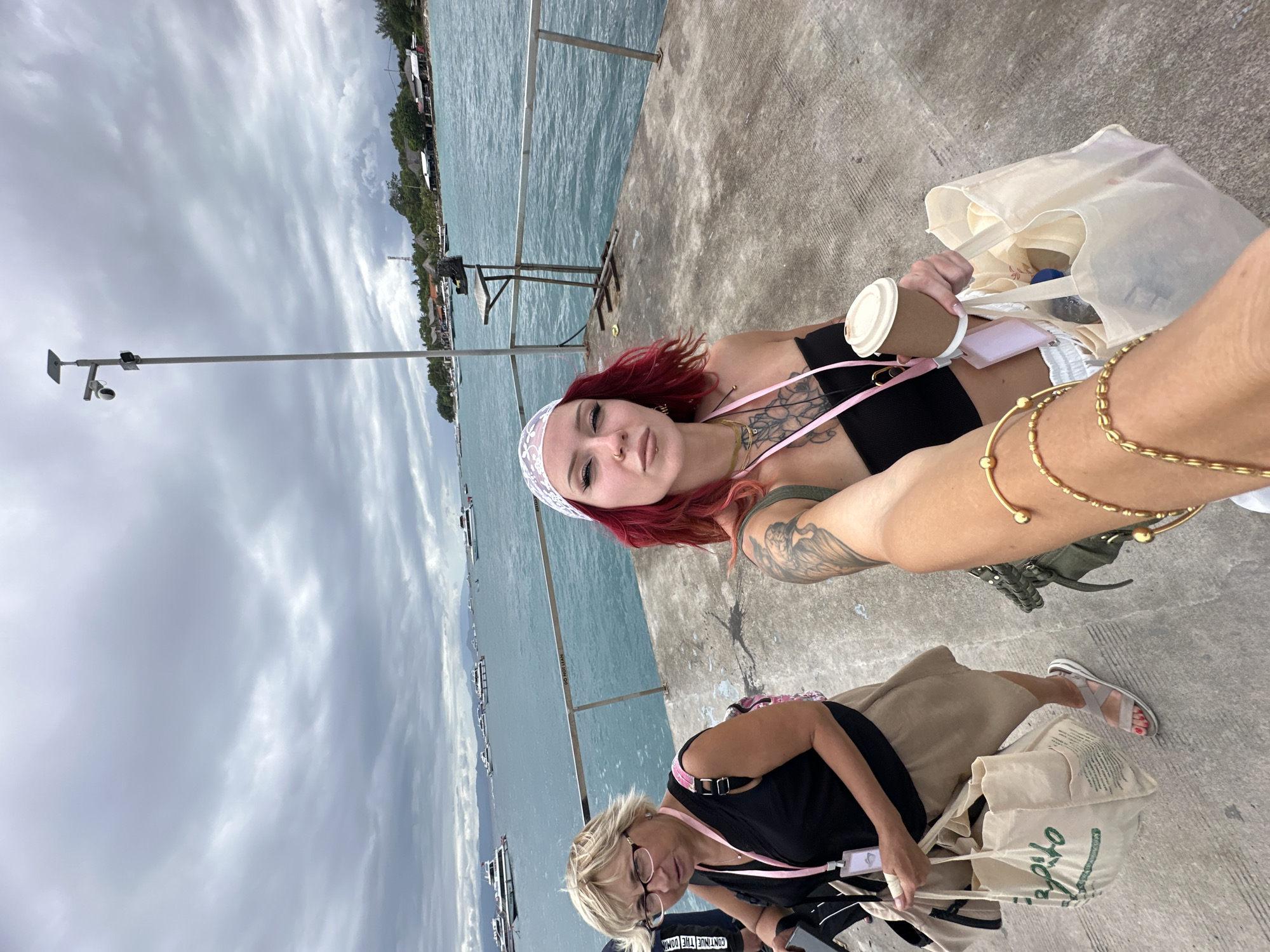 Two women are on a concrete pier in Kampung Toyapakeh, Indonesia. One woman has red hair, a white headband, and tattoos on her arm. She wears a black top and gold bracelets. She is holding a coffee cup and a white bag. The other woman is blonde and wears glasses, a black top, a khaki skirt, and sandals. She also holds a white bag. The sea is in the background with some boats and the sky is cloudy.