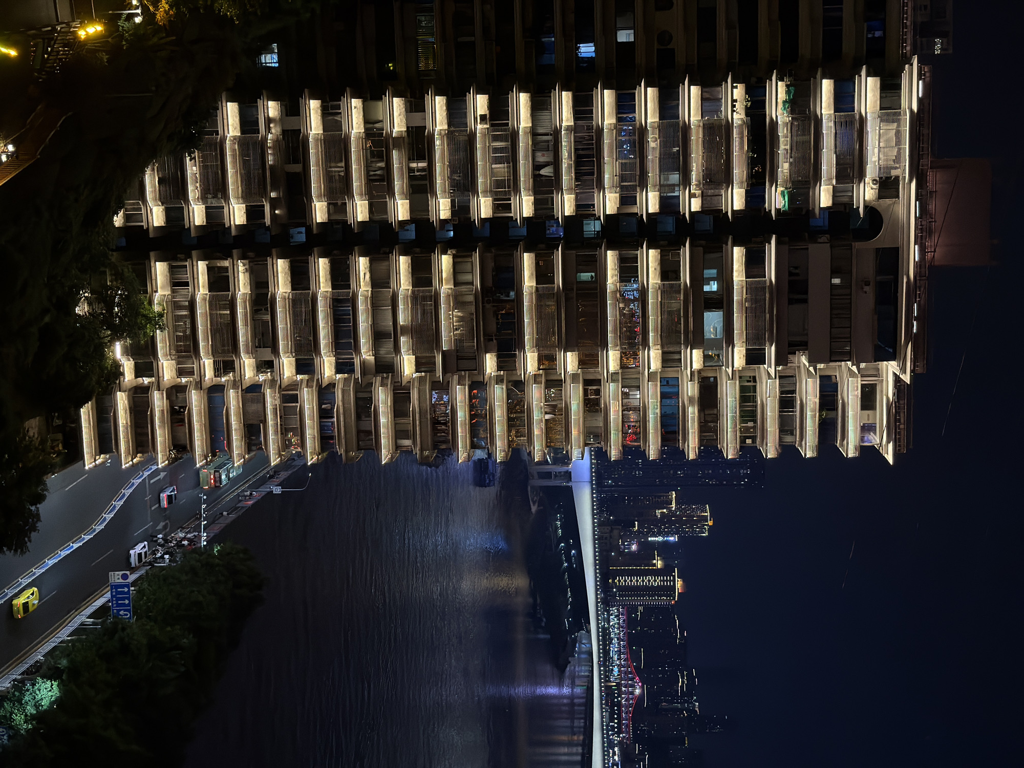 This image captures a nighttime view of a modern, multi-story building illuminated by warm lights along its balconies. The building is situated in Yuzhong District, China, as evidenced by the prominent Chongqing skyline and Yangtze River in the background. The river's dark water reflects the city lights and a brightly lit bridge, suggesting a bustling urban environment. A street runs alongside the building, with a few cars, including a yellow taxi, visible, indicating ongoing traffic. Pedestrians can be seen gathered on the sidewalk. Trees and greenery surround the base of the building and line the street, adding a touch of nature to the cityscape. The overall scene depicts a lively urban nightscape with architectural highlights and the presence of human activity.