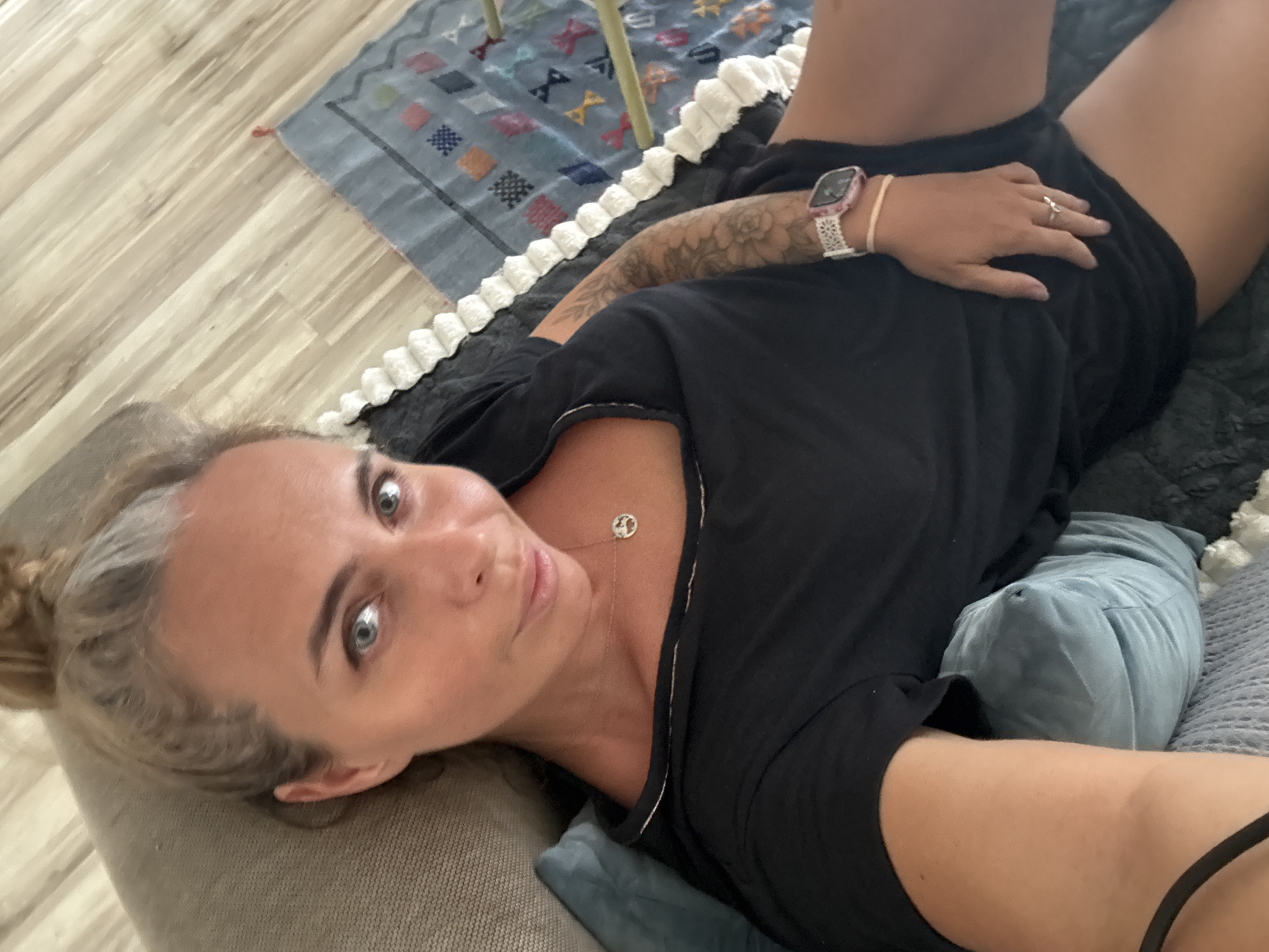 A woman is lying on a couch in a room that appears to be in Ko Samui District, Thailand. The woman has blonde hair, blue eyes, and is wearing a black t-shirt and black shorts. She is also wearing a watch on her left wrist and a ring on her right ring finger. There is a blue pillow and a gray blanket on the couch with her. In the background, there is a rug on the floor and a part of a chair. The room has light wooden floors and appears to be well-lit.