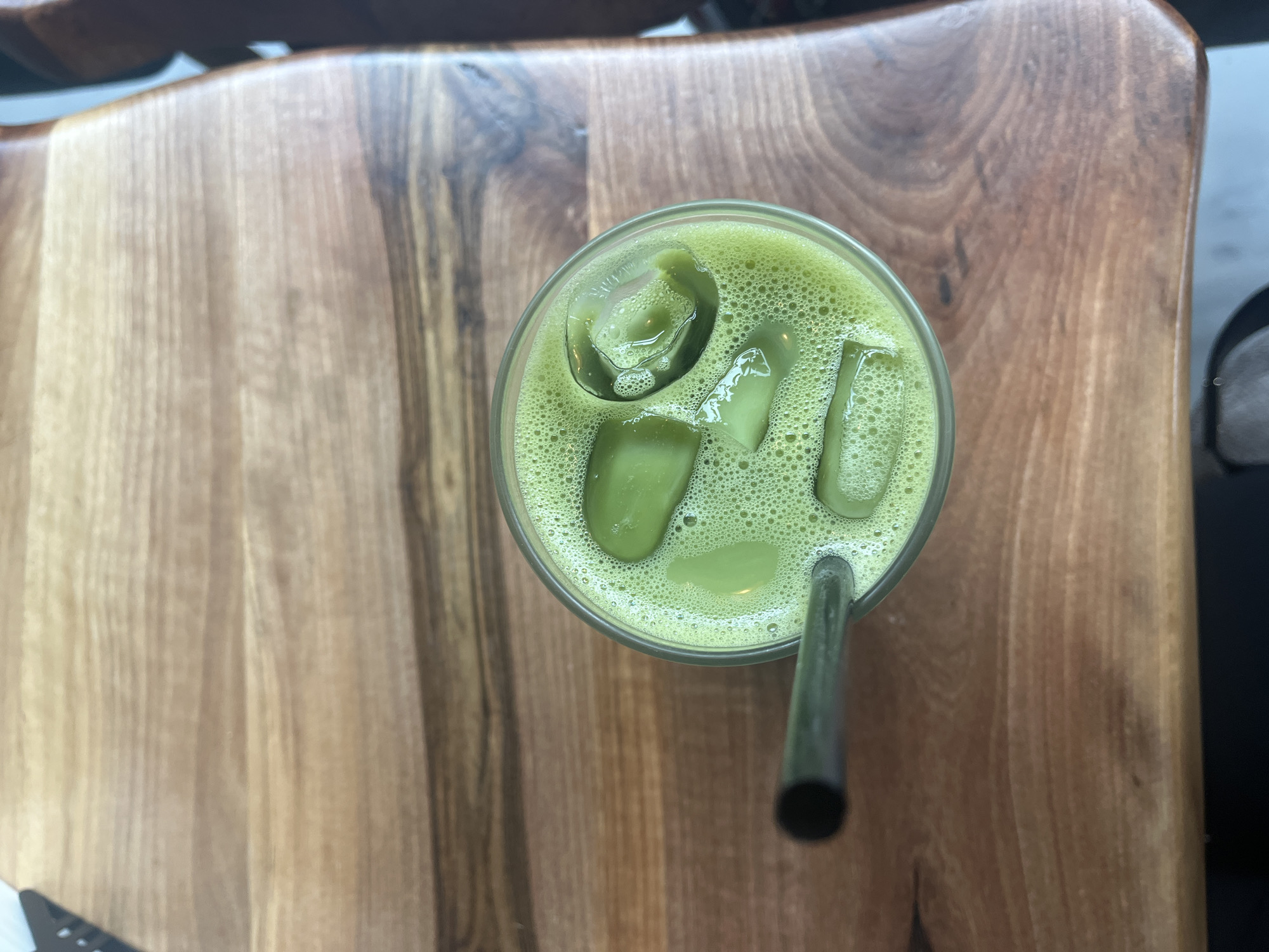 A top-down, medium-shot depicts a glass of vibrant green liquid, likely a matcha-based beverage, containing ice cubes and a dark metal straw. The glass rests on a wooden surface displaying varied grain patterns and shades, suggesting it might be a tabletop or a serving tray. The setting seems to be indoors, possibly a café or restaurant. The scene has a neutral, natural lighting scheme. The photo was taken in Plovdiv, Bulgaria.