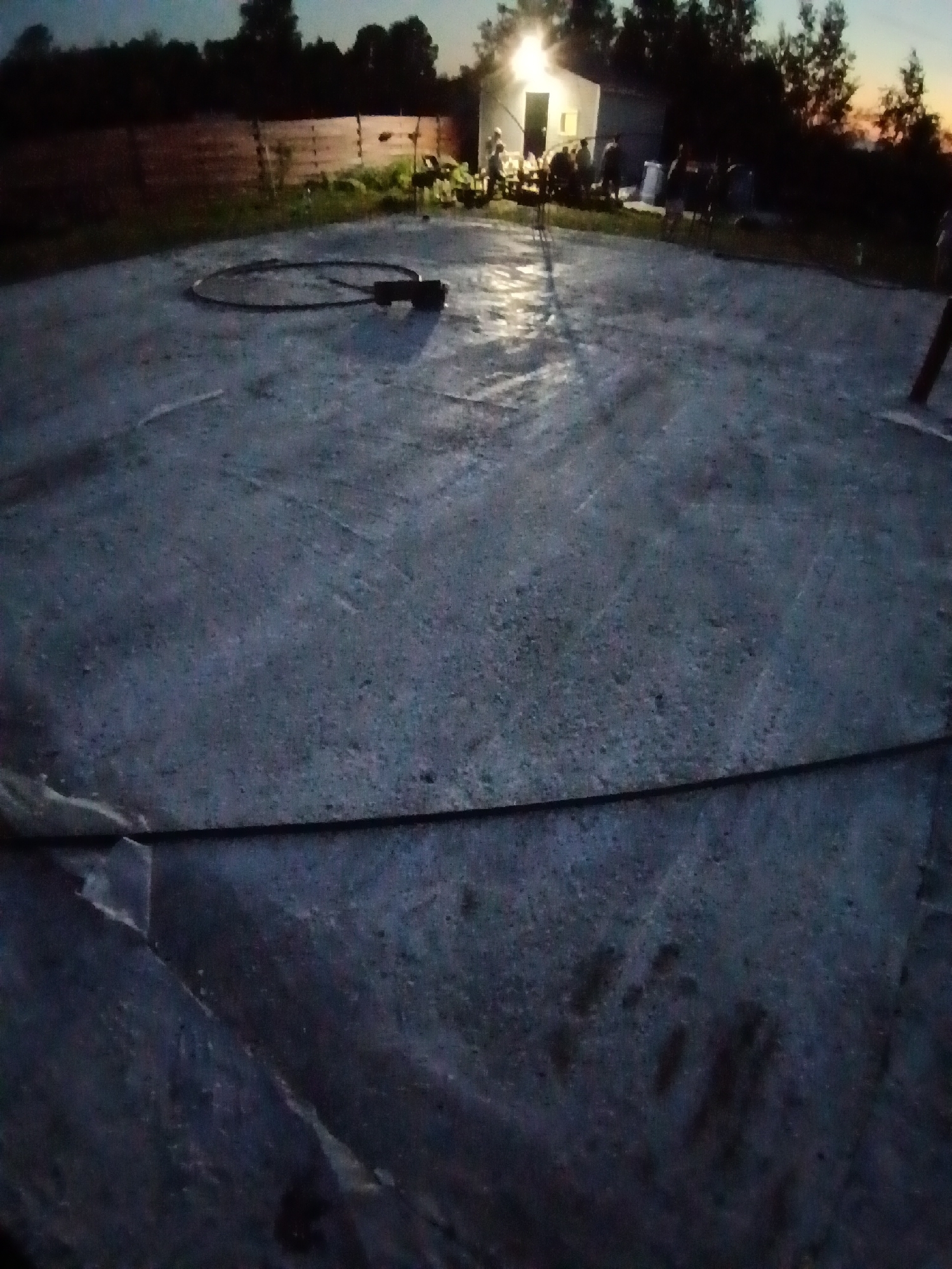 The image depicts an outdoor scene at dusk, possibly in Amursk, Russia, though this location is not explicitly confirmed. In the foreground, a large, grey, textured surface, likely concrete, occupies most of the frame. A black, coiled hose with a black object attached to one end lies on this surface in a circular pattern. In the background, several silhouetted figures are gathered around a brightly lit, white structure that appears to be a building or a large tent. Trees and a wooden fence are also visible in the dimly lit environment. The overall lighting suggests it is twilight or early evening, with artificial light emanating from the structure in the background.