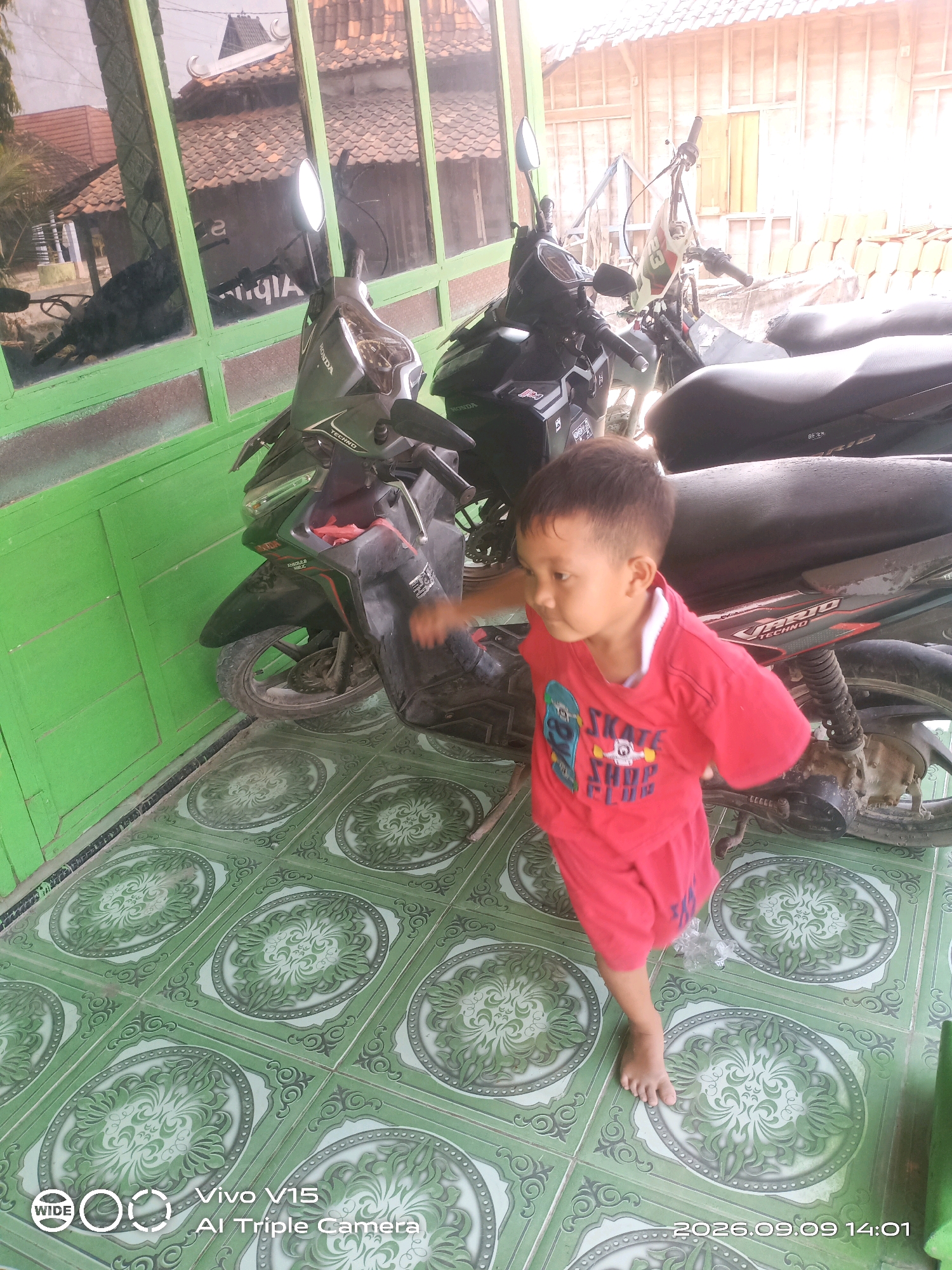 A young boy wearing a red t-shirt with a skateboard graphic and red shorts stands on a green tiled floor. He is looking to his left, with his right arm extended. Behind him are several parked motorcycles. The scene appears to be an outdoor area, possibly a porch or driveway, with a green wall to the left and a wooden structure with a tiled roof in the background. The image is taken from a medium-close-up perspective, focusing on the boy and the motorcycles. The time stamp at the bottom right indicates the date as 2026.09.09 and the time as 14:01.
