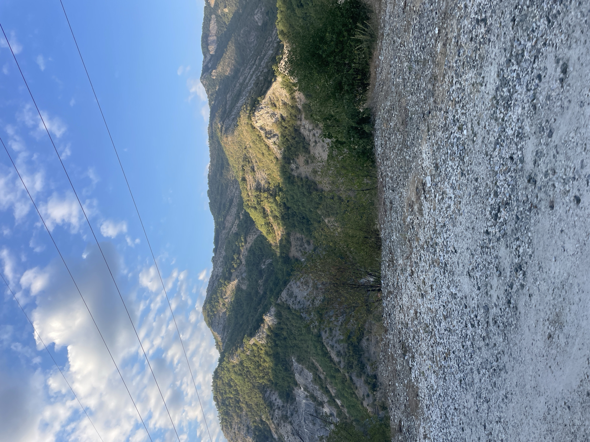 The image depicts a mountainous landscape with scattered vegetation under a bright blue sky with some white clouds. In the foreground, a gravelly and dusty ground surface is visible. Three parallel power lines stretch horizontally across the upper portion of the image, above the mountains. The mountains themselves are covered in green trees and rocky patches, with some areas illuminated by sunlight and others in shadow. The scene appears to be a natural outdoor environment.