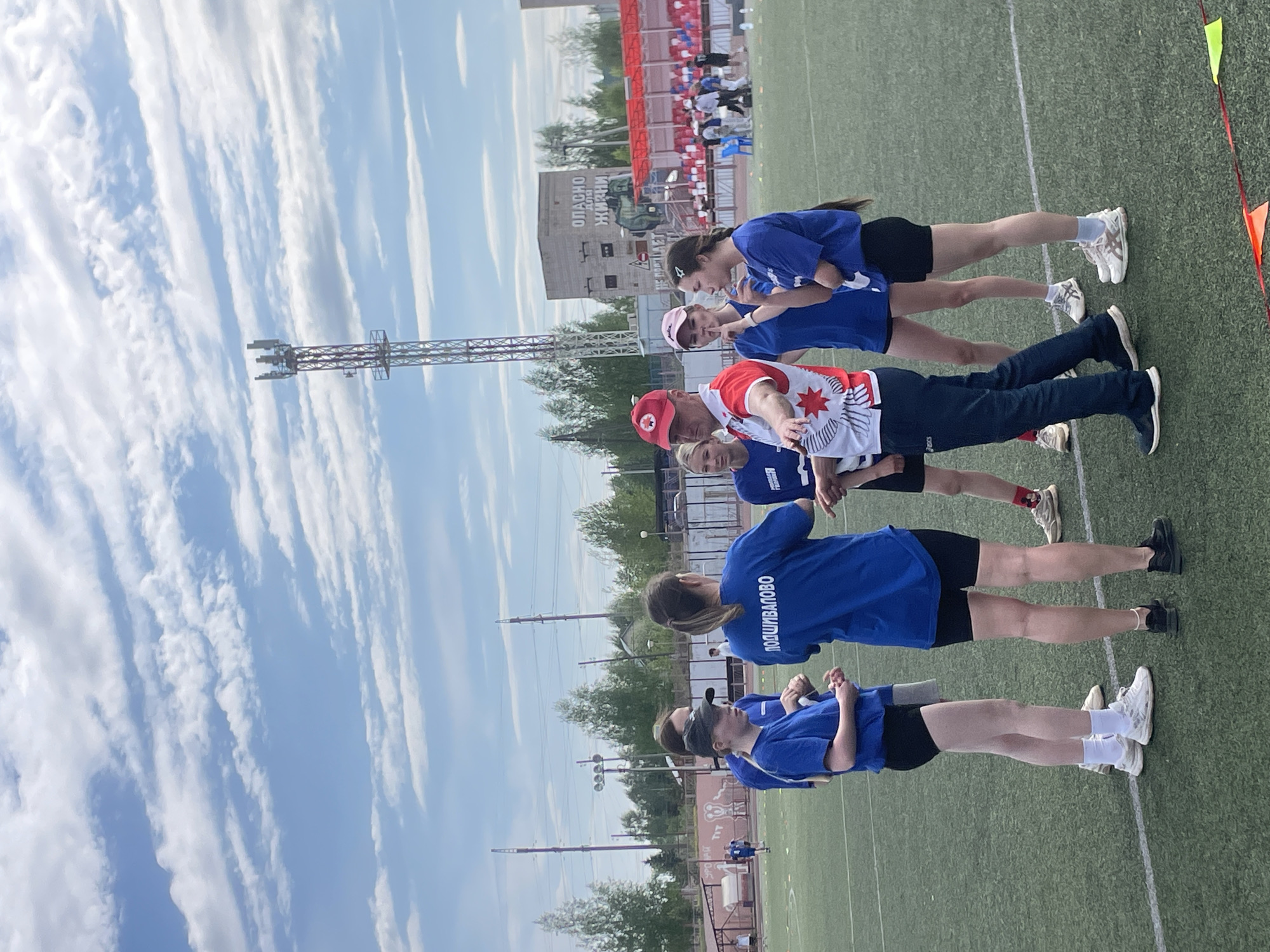A medium shot of a group of individuals on a field in Zav'yalovo, Russia. They are standing in a row. The individuals are dressed in blue shirts, black shorts, and various types of sneakers. One individual is wearing a red cap and a jacket with a white design and a red star. The environment includes a field, a building in the background with signage in Russian, a tall metal structure, and a sky with wispy clouds.