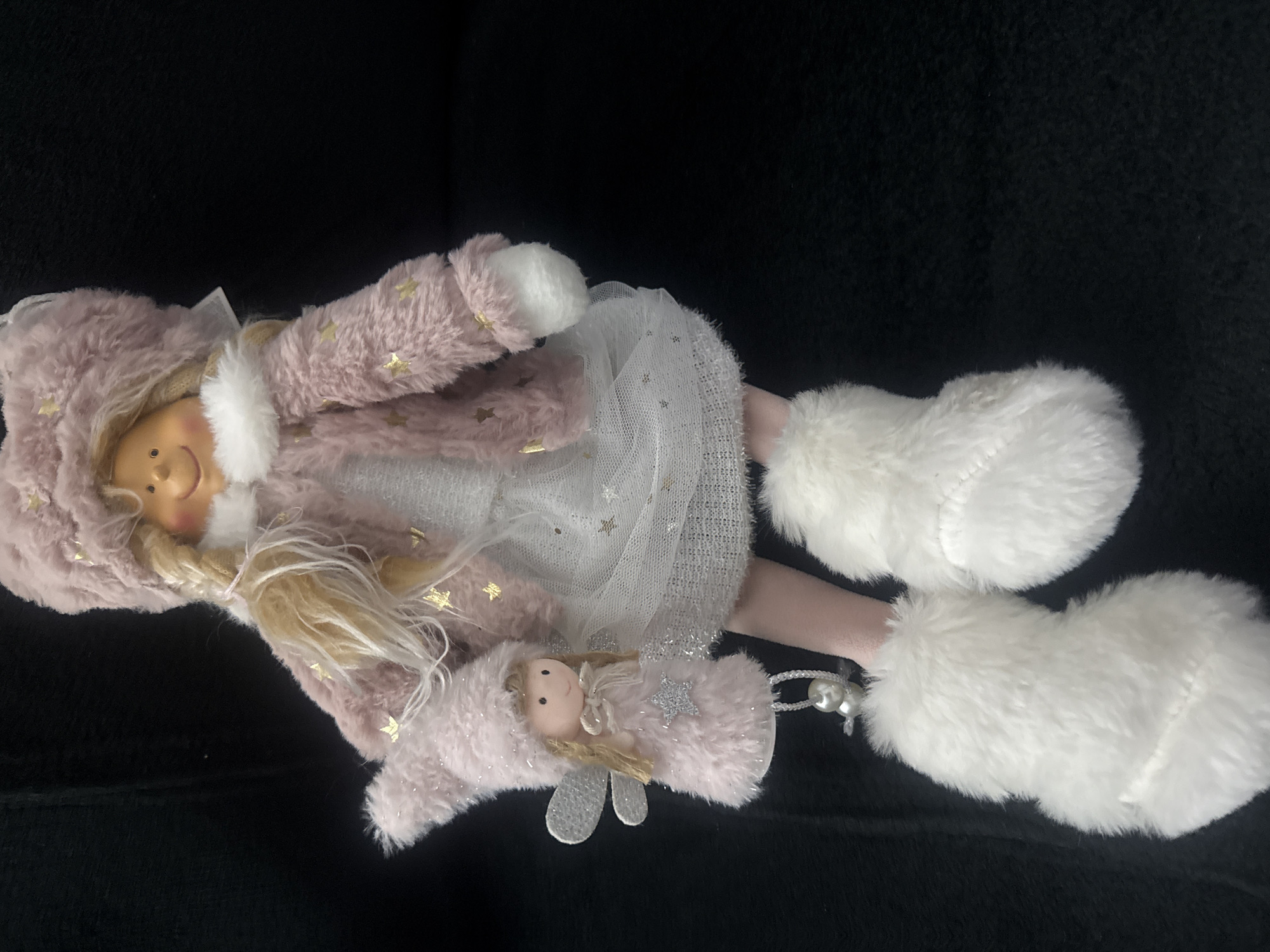 A doll is positioned against a black backdrop. The doll is attired in a pink, fur-like coat adorned with gold stars, paired with a white, netted dress. It features blonde hair and white, fur-like boots. The doll holds a smaller doll, similarly clad in pink fur and featuring silver wings. The scene appears to be set in Unknown, Unknown.