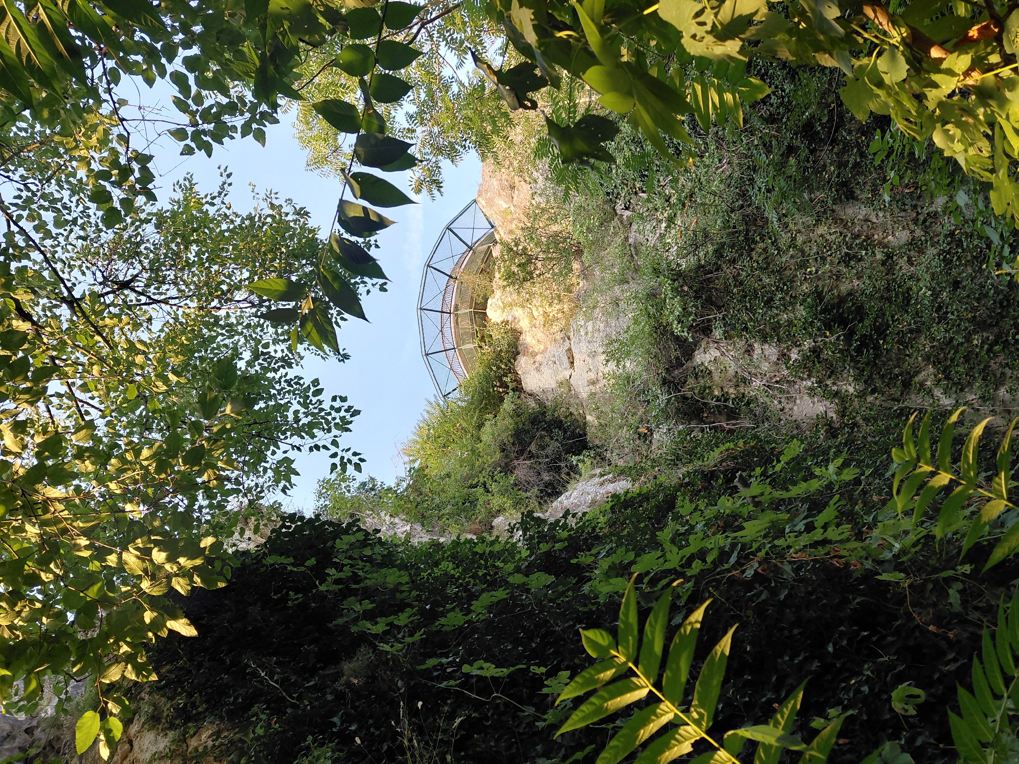 The image depicts a view upwards towards a cliff face partially covered in ivy and other vegetation. At the top of the cliff, a circular, open-air structure with a metal railing and a netted enclosure is visible. This structure appears to be a viewing platform or observation deck. The scene is framed by green leaves and branches from trees in the foreground, with a clear blue sky visible in the background. The lighting suggests it is daytime, possibly late afternoon given the warm tones on the rock. This location is the Pleven Lookout (also known as the "Panorama" or "Pleven Key"), situated in the city of Pleven, Bulgaria, and is part of the Pleven's Liberation panorama complex.