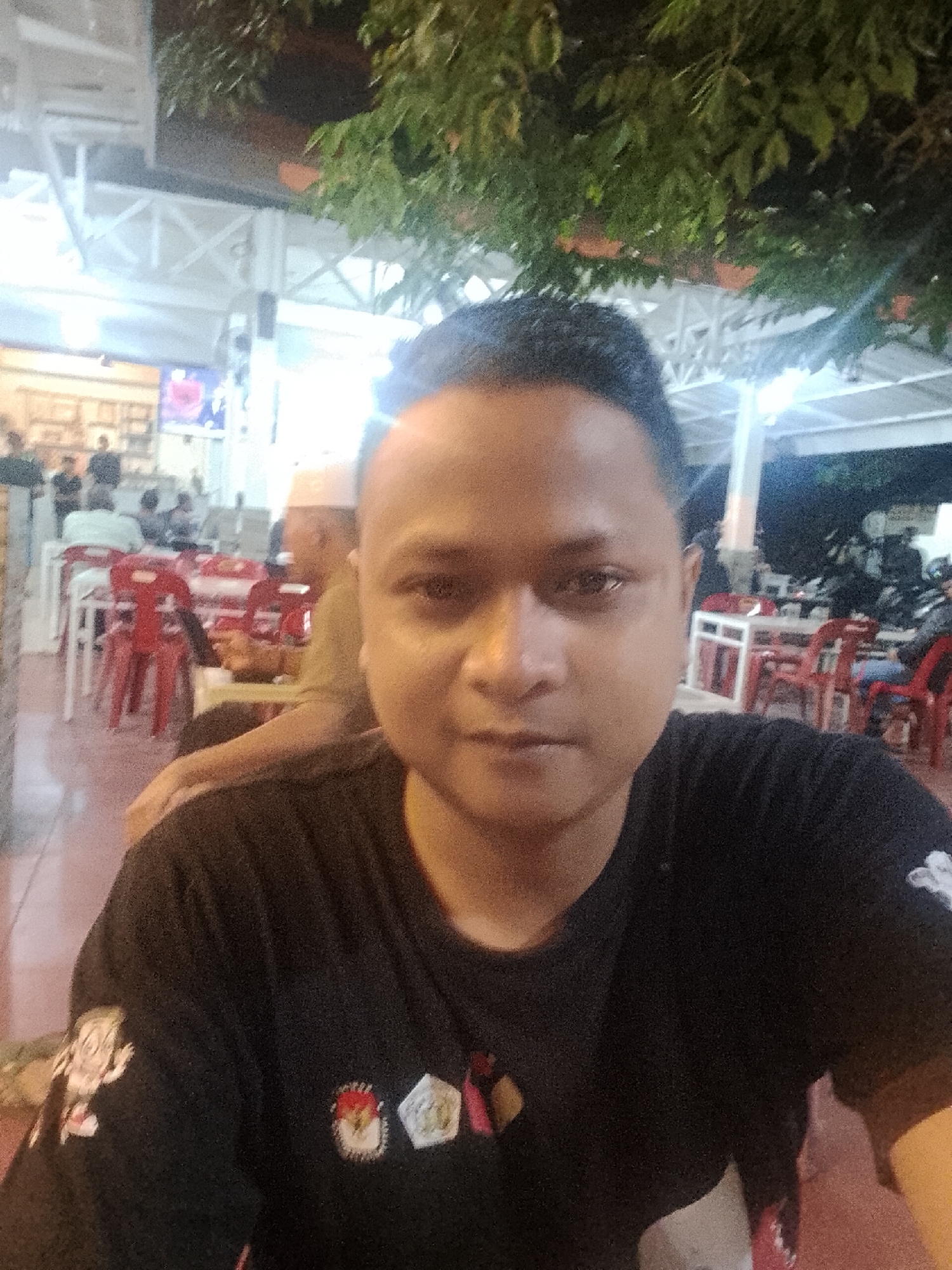 A man is captured in a selfie-style shot, looking directly at the camera with a neutral expression. He is wearing a black t-shirt featuring embroidered patches; one on his left sleeve depicts a cartoon character, while on his chest, there are at least two distinct badges. One of these is a pentagonal emblem clearly identifiable as the Garuda Pancasila, the national symbol of Indonesia, confirming the location context of Lhokseumawe, Indonesia. Another patch next to it is circular with red and white elements.
The scene is set at night, indicated by the dark background and prominent artificial lighting. Overhead, tree foliage is visible, along with white structural beams and supports of a building or canopy. In the background, a casual outdoor or semi-outdoor dining area is visible, populated by other individuals. A man wearing a white cap (kufi) is partially visible in the mid-ground. Red plastic chairs and white tables are scattered throughout, occupied by several blurred figures who appear to be socializing or dining. Bright light sources illuminate parts of the background, and faint shapes resembling parked motorcycles are discernible in the far right.