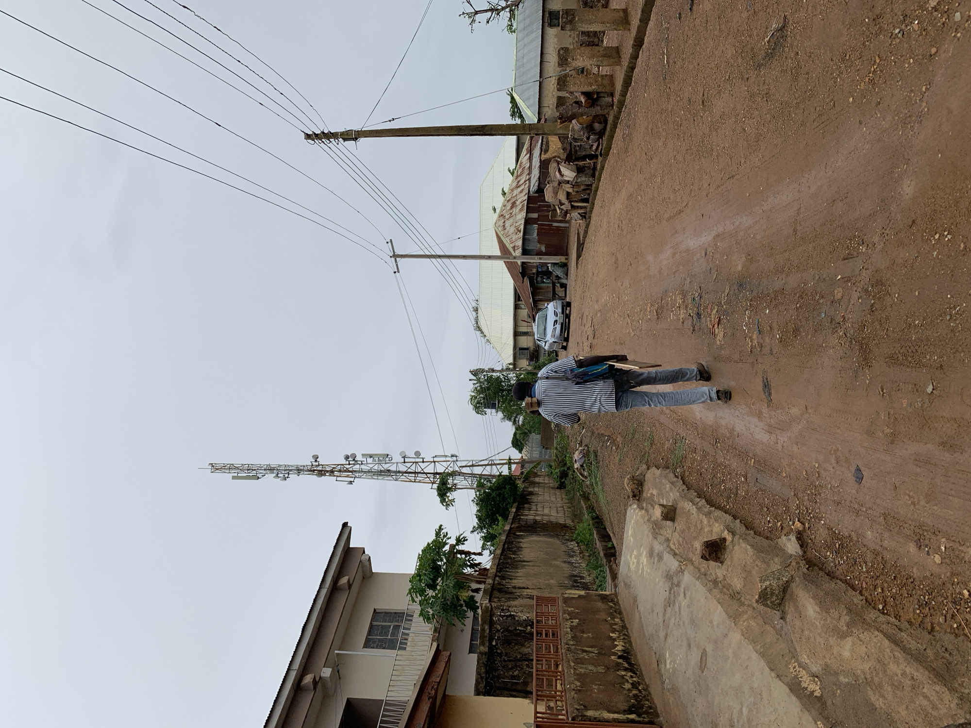 The image depicts a dirt road in Rafin Par, Nigeria, on an overcast day. A person is walking away from the camera on the right side of the road, wearing a striped shirt and jeans, with a bag slung over their shoulder. To their left, on the other side of the road, is a house with a metal fence and a cell tower. Utility poles with power lines run along the road above. Further down the road is a building with a corrugated metal roof, a car, and a collection of materials stacked outside. The road surface appears uneven and muddy.