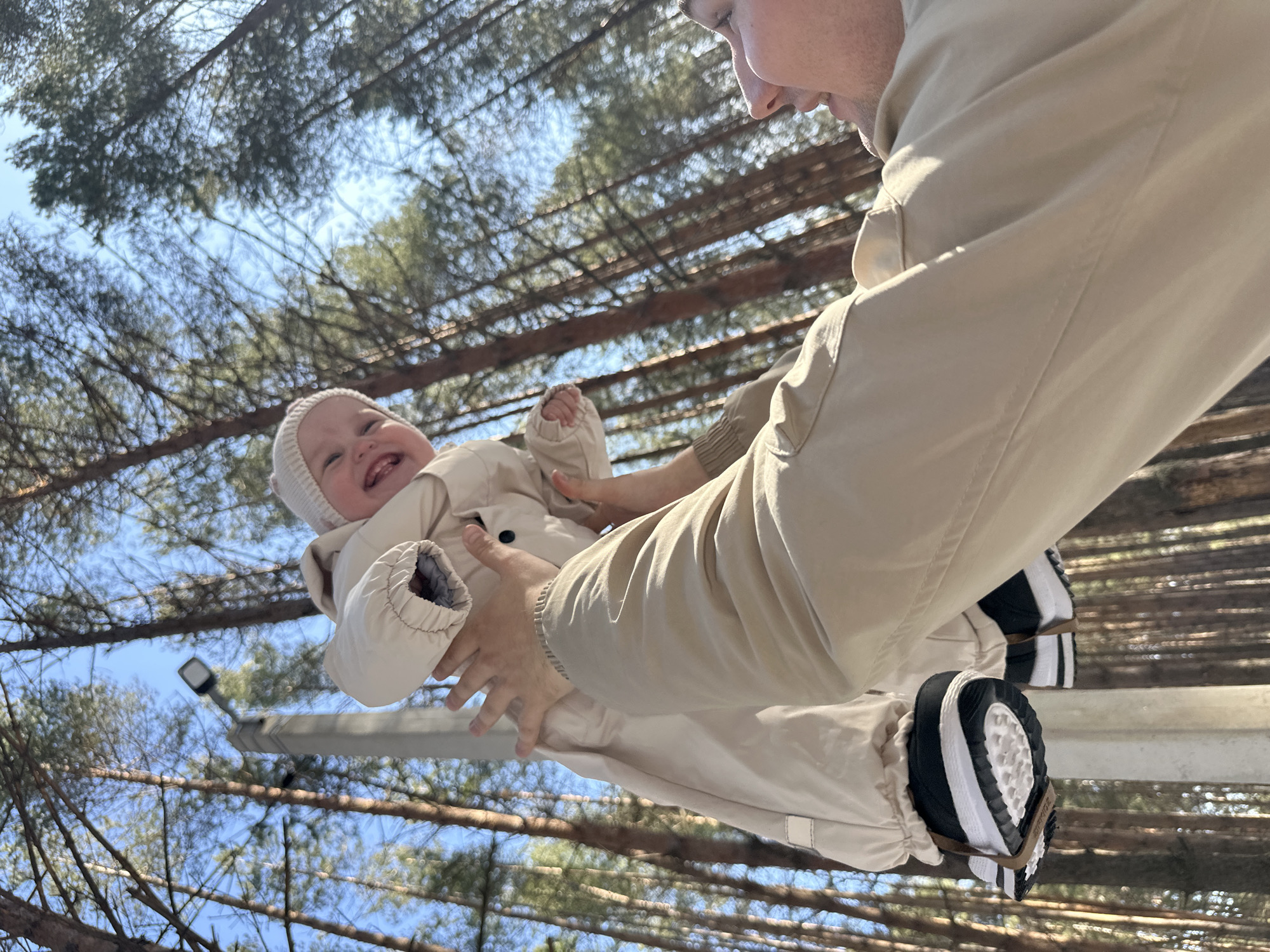 The image depicts an outdoor scene in the city of Chaikovsky, Russia. A man, partially visible in the lower right, wears a light tan long-sleeved shirt and looks upward with a slight smile. He is holding an infant aloft in the center-left portion of the frame. The infant wears a light beige padded snowsuit, a white knitted beanie, and black and white shoes with prominent white and black soles. The infant is smiling broadly, facing the camera, with an open mouth and visible teeth. The man's hands support the infant's torso. In the background, numerous tall conifer trees stand against a clear blue sky. A light grey concrete pole with a dark rectangular light fixture at its top extends vertically behind the infant's lower body and feet.