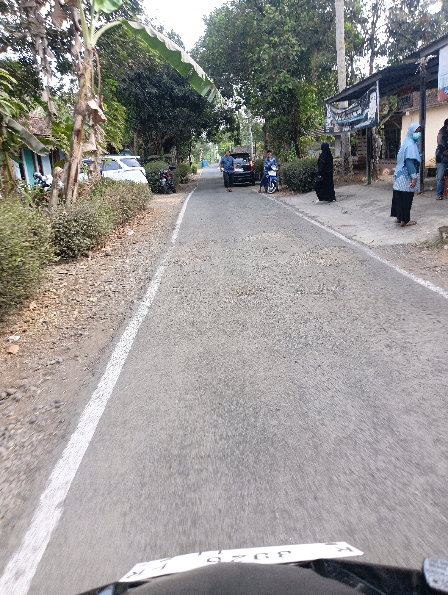 The image captures a street scene in Majatengah, Indonesia. A paved road with a white lane marker runs through the frame. On the left side of the road, there is a white minivan parked near a house with a tiled roof and a blue door. A motorcycle is also parked nearby. A lush green bush borders the road on the left. Further down the road on the right, a black car is stopped, and two men, one in a blue shirt and the other in a patterned blue shirt, are standing near it. A blue motorcycle is also present. Several people are standing on the sidewalk on the right side of the road. One person, wearing a black hijab and abaya, is standing, while another individual in a light blue hijab and a striped top with black pants is also standing. A third person, in a blue shirt and black pants, is visible further down. The environment is characterized by trees lining the sides of the road, with some buildings and structures visible in the background. The overall scene appears to be a typical day in a residential or semi-rural area of Majatengah.