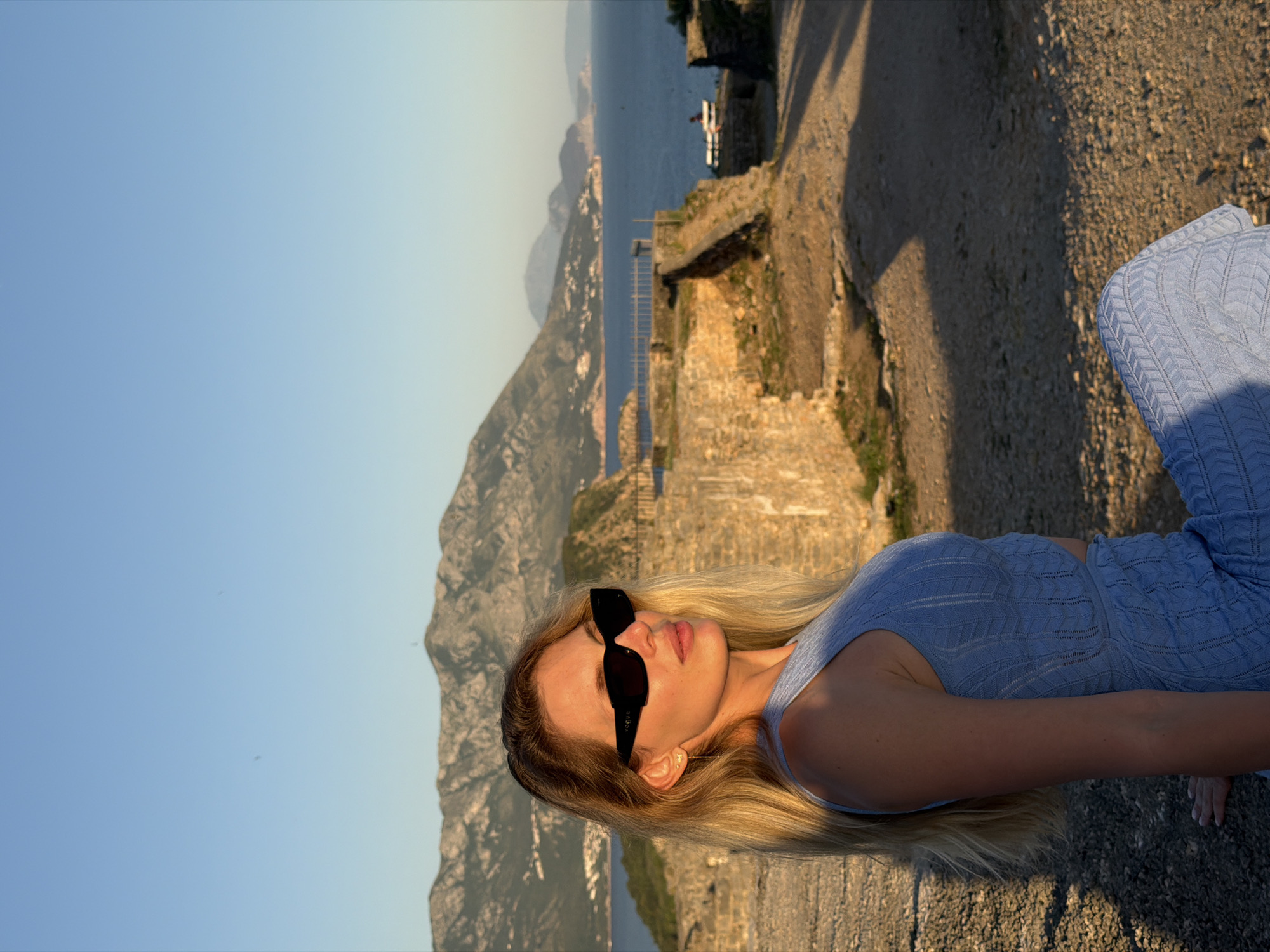 The image presents an outdoor scene at golden hour, characterized by warm, directional light and strong shadows. The setting is an elevated coastal viewpoint featuring ancient stone ruins overlooking the Adriatic Sea, consistent with locations around Budva, Montenegro.
The central subject is a young adult female with long blonde hair, seated on a low, rough stone wall in the foreground. She wears black, rectangular-framed sunglasses with "VOGUE" printed on the left temple. Her left ear displays three small gold stud earrings and a thin gold chain earring dangling from the lowest piercing. She is dressed in a light blue knitted two-piece outfit, consisting of a sleeveless crop top and a matching skirt or trousers, both featuring a patterned knit design. Her body is angled slightly to the left, with her head turned to the right, presenting her right profile and the left side of her face illuminated by the setting sun. Her gaze is directed towards the right side of the frame.
Behind her, weathered stone ruins and a dirt path with visible shadows occupy the mid-ground. Further in the background, a calm, deep blue body of water, the Adriatic Sea, stretches towards the horizon. Distant, rugged mountains rise from the left side of the frame, with visible coastal settlements along their base. A smaller, green landmass or island is present in the mid-right background. A lone individual is discernible seated on a white bench near the water's edge on the far right, amidst the ruins. The sky above is clear and light blue, with two small, dark specks visible.