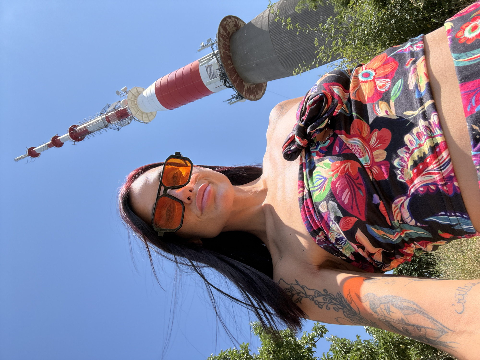 A woman with dark hair and bright pink streaks, wearing large orange-tinted sunglasses and a colorful floral strapless top, stands outside on a sunny day. She is positioned in the foreground, looking upwards towards a tall, red and white striped telecommunications tower that dominates the background. The tower is situated in the city of Pleven, Bulgaria. Green trees and foliage are visible on the left and right sides of the frame, suggesting an outdoor, elevated location. The woman has tattoos on her left arm. The image is a low-angle, wide shot, likely a selfie.