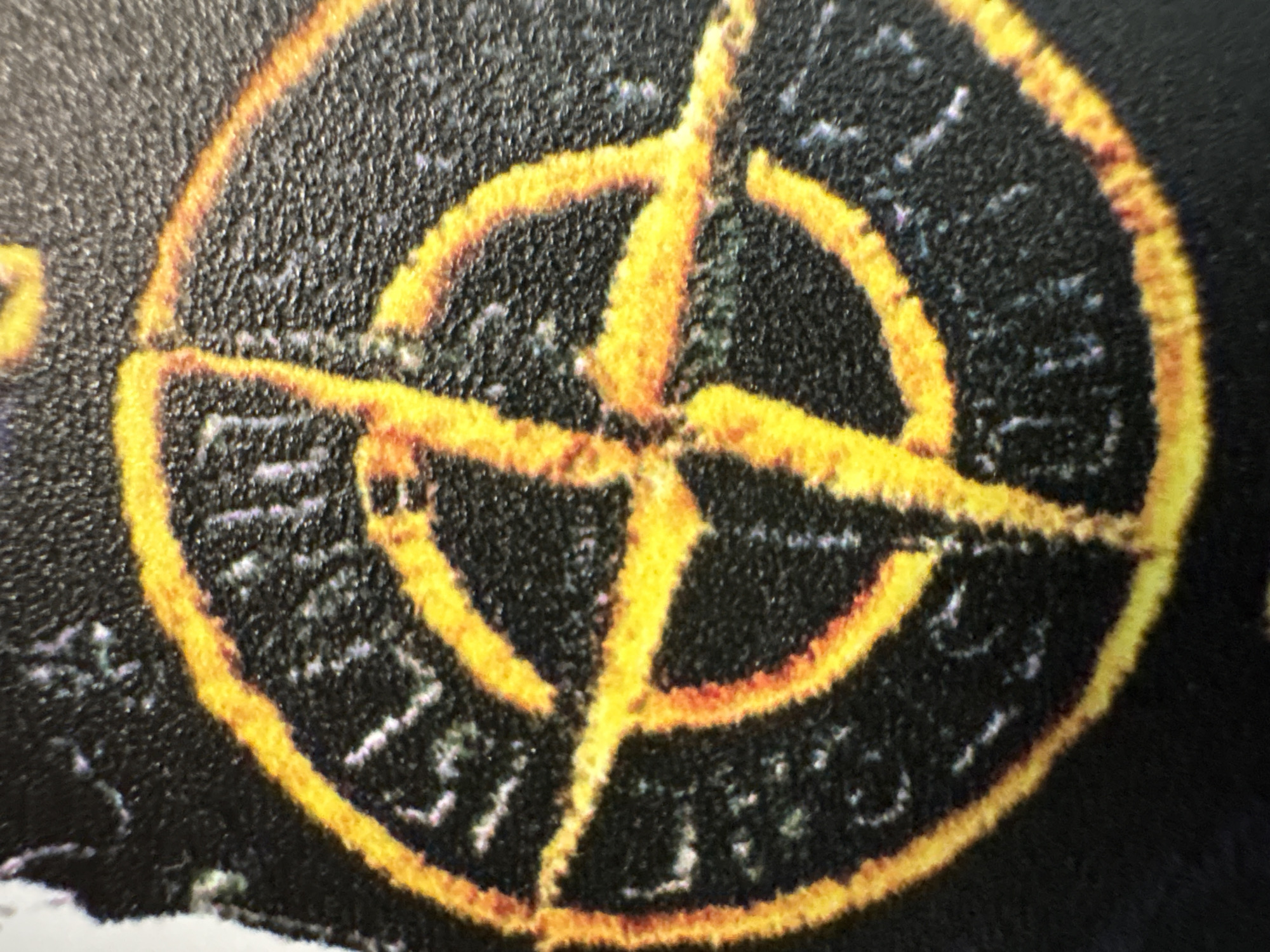 The image displays a close-up view of a black fabric patch with a yellow and orange compass-like symbol. The symbol features a circular outer edge with radiating lines extending inwards, forming a four-pointed star. Some text is visible within the symbol, appearing in a faded white or light gray color against the black background. The texture of the fabric is rough and granular. The image does not depict any identifiable urban or natural environments, and therefore, no specific location in Dnipro, Ukraine, can be determined. There are no people, animals, or discernible actions or interactions present in the image.